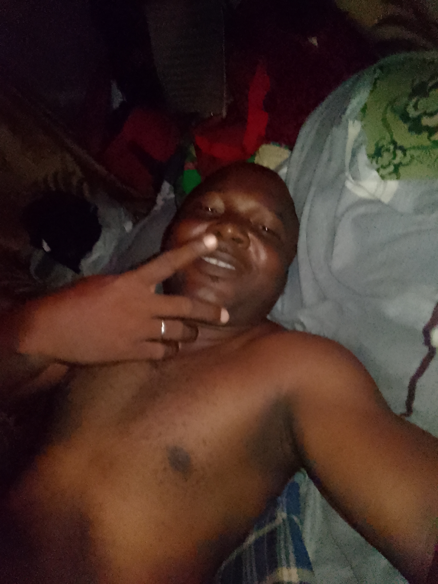 A shirtless, dark-skinned man lies on a gray patterned cloth with a ring on the fourth finger of his left hand, with that hand making a peace sign near his face. The surrounding area includes what appears to be rumpled clothing and bedding of various colors. The location appears to be inside a building, though further details are undiscernible.