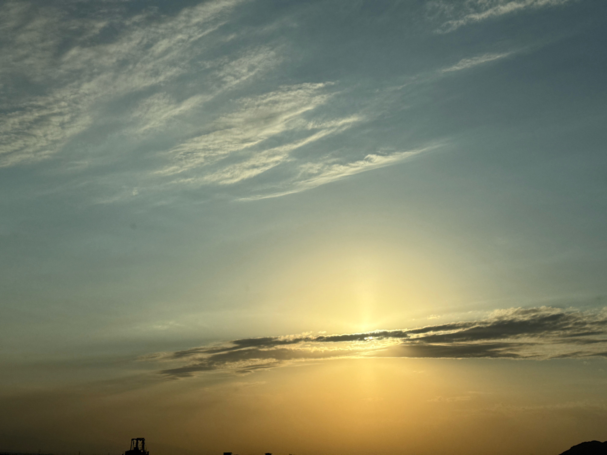 The image shows a wide sky with wispy clouds, transitioning from a pale blue at the top to a warm yellow and orange gradient near the horizon. The sun is partially obscured by a band of clouds, casting bright rays through them. In the lower portion of the image, a dark silhouette of what appears to be heavy machinery, possibly a forklift or construction equipment, is visible against the brighter sky. The ground is mostly obscured by shadow and atmospheric haze, but faint shapes suggest distant structures or terrain. The scene is characteristic of a sunset or sunrise over an open area, possibly industrial or agricultural, near the city of Chüy, Kyrgyzstan. There are no people or discernible interactions visible.