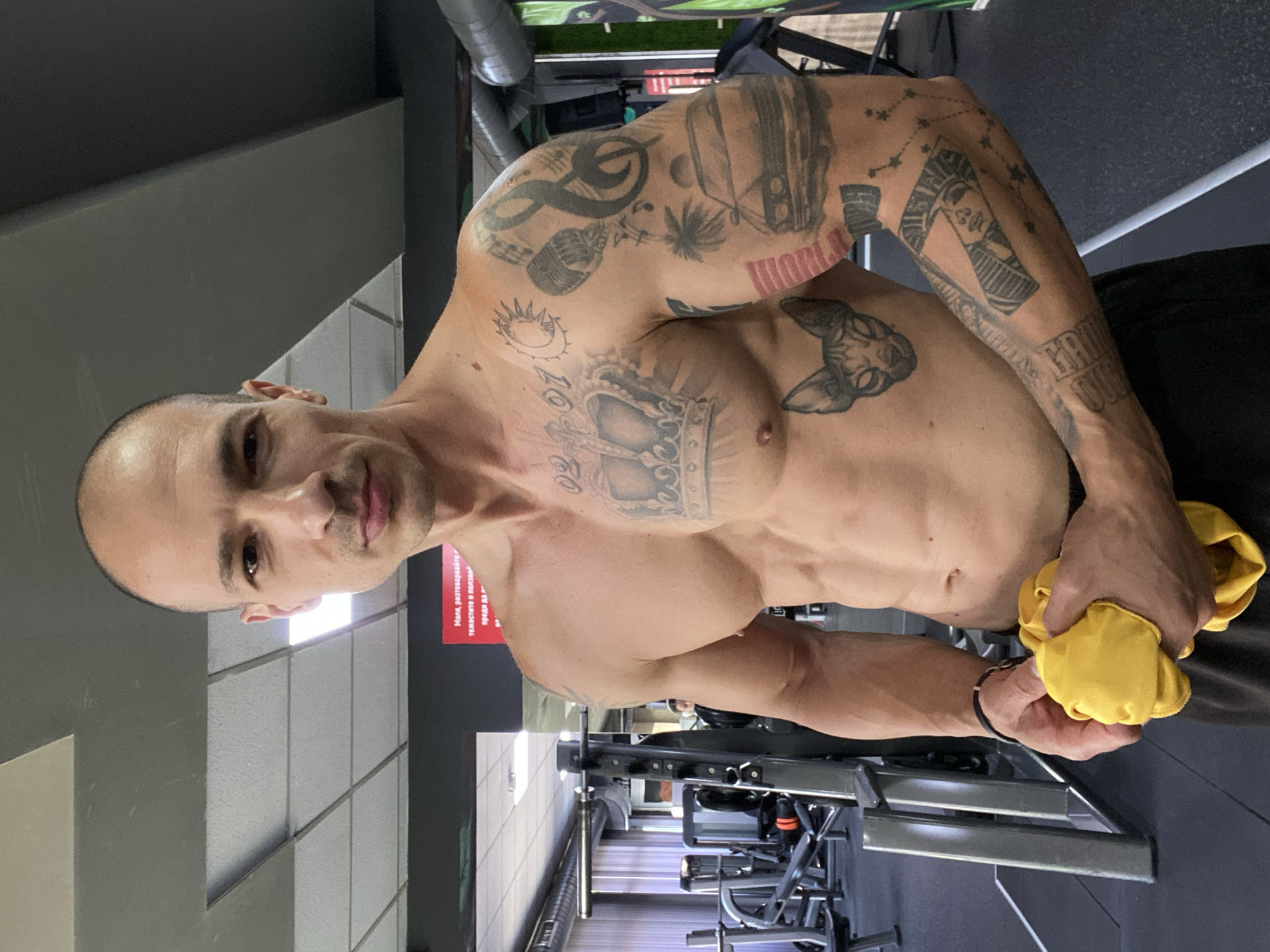 A bald man with visible musculature and tattoos stands in a gym. He is shirtless, wearing dark shorts, and holding a yellow towel in his hands. The background shows gym equipment, including weight machines and racks. A red sign with Cyrillic text is visible on the wall. The man's tattoos include a crown on his chest, a cat's face on his abdomen, and various designs on his arms, such as a car, a microphone, and musical notes. The lighting suggests an indoor environment, likely during the day. The location is identified as a gym in Pleven, Bulgaria.