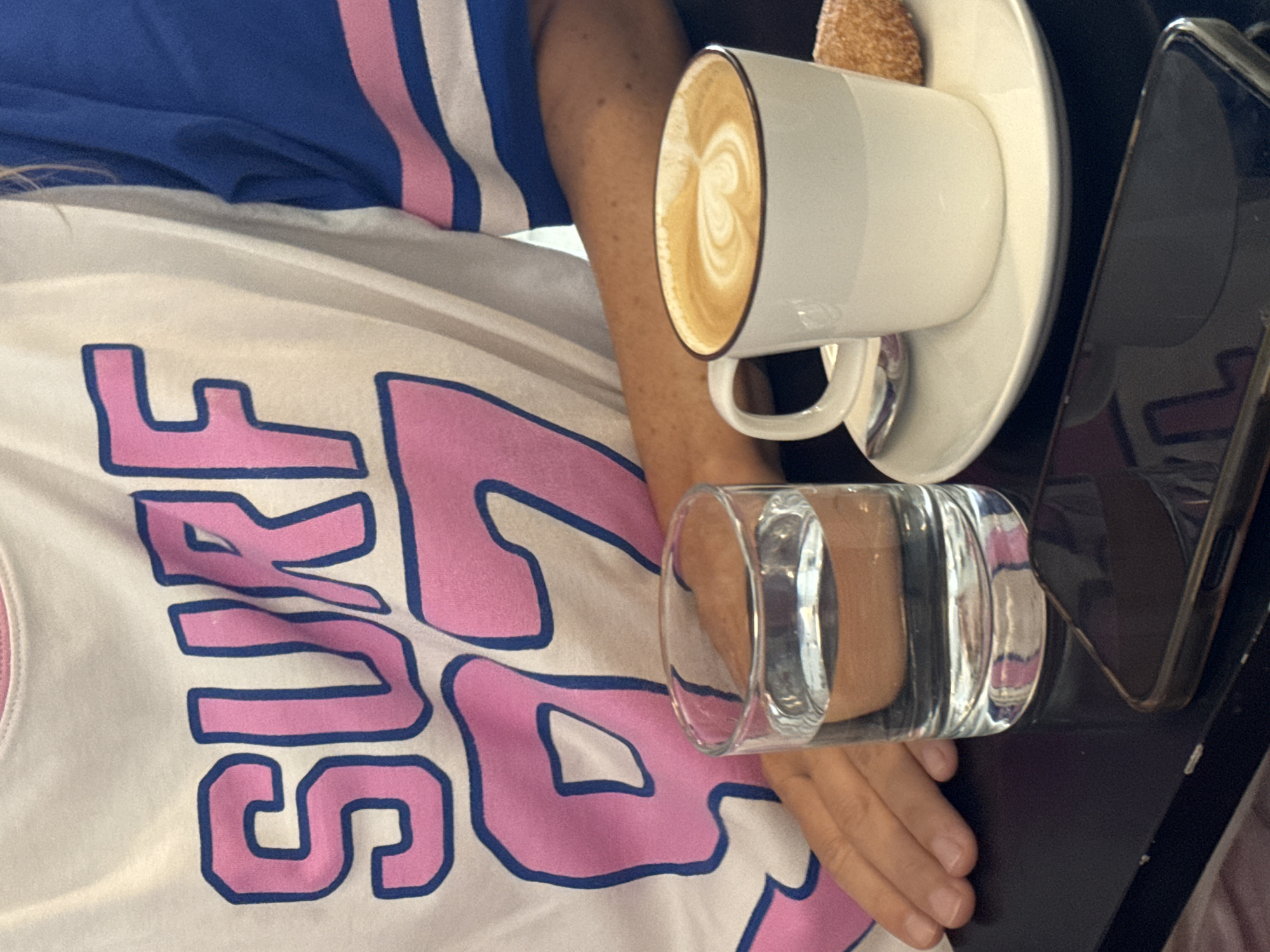 A person in a white t-shirt with pink and blue "SURF" lettering is sitting at a dark table, possibly in a cafe in Kuta Selatan, Indonesia. On the table is a cup of coffee with latte art on a saucer, a small pastry, a glass of water, and a smartphone. The person's hand is visible near the glass of water. The scene is captured from a top-down perspective.