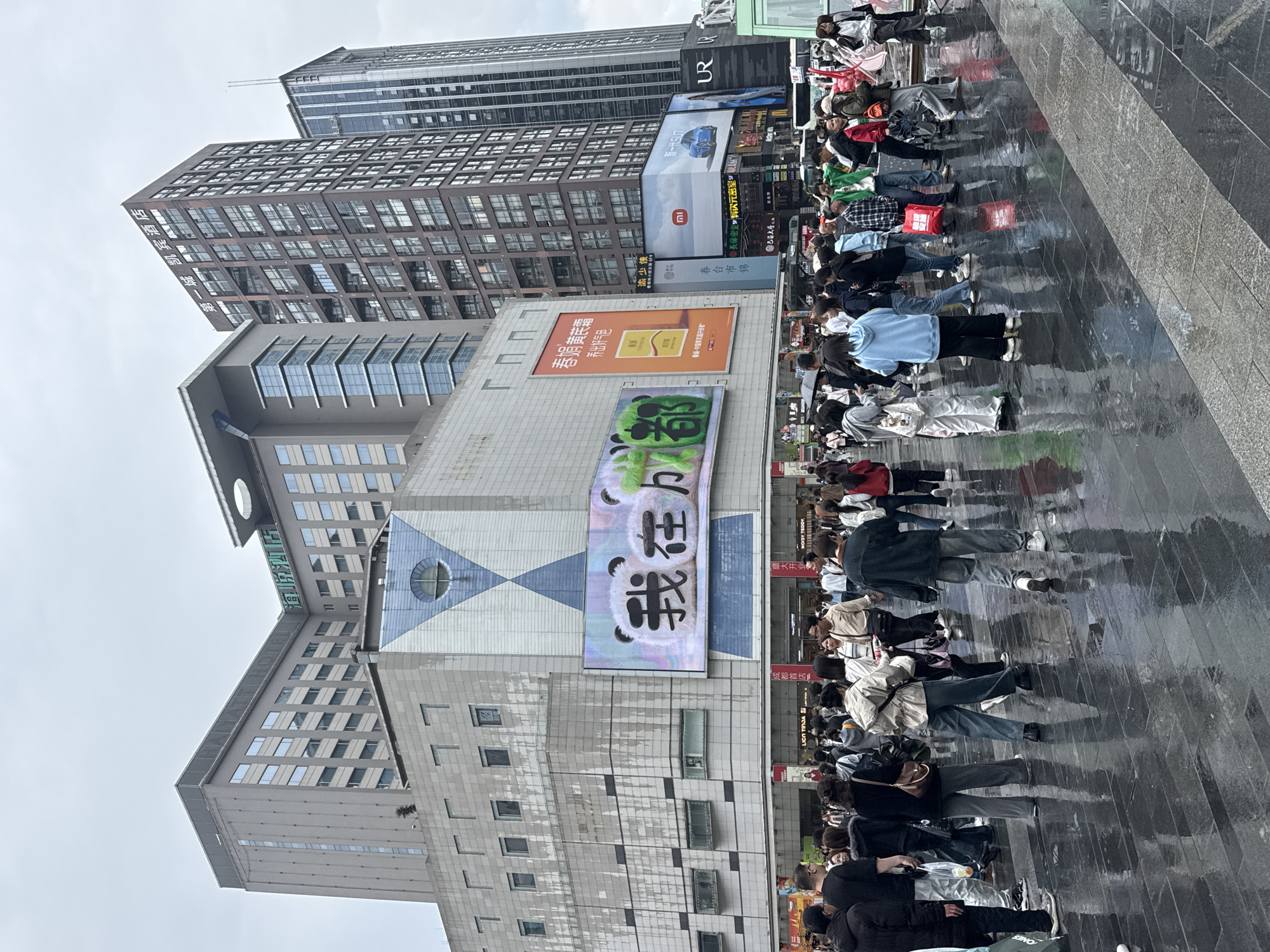 The image captures a street scene in Jinjiang District, China, with a large crowd of people filling a wet paved area. Several high-rise buildings dominate the background, featuring a mix of architectural styles and commercial advertisements. One building displays a large electronic billboard with panda imagery and Chinese characters. The street is lined with shops and various commercial signs. The ground is reflective, indicating recent rain. The crowd appears to be a mix of locals and tourists, engaged in shopping and leisure activities.