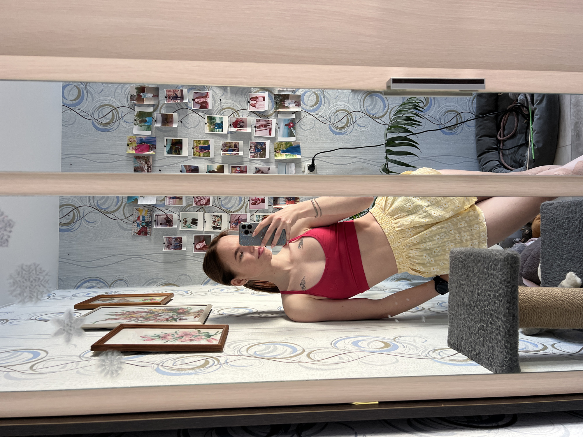 An interior scene captured through a full-length mirror, revealing a young adult female taking a selfie. The woman, with brown hair tied back in a ponytail, wears a red sports bra and yellow patterned shorts. She is positioned with her right shoulder leaning against a wall, holding a smartphone with her right hand to capture her reflection. Two small tattoos are visible on her chest, below her collarbones.
The environment reflected in the mirror is an indoor residential space, likely in the city of Chaikovsky, Russia. The wall to the left of the woman features light-colored wallpaper with a recurring blue and brown circular pattern. Three framed pictures, depicting floral designs, are mounted on this wall. Three white, snowflake-shaped decorations are also affixed to this wall. The wall to the right, also with the same patterned wallpaper, is densely covered with numerous small photographs pinned to its surface, forming a collage. A black power cord is visible trailing down this wall from a white electrical outlet. A tall, dark green houseplant is visible in the lower right background, behind a dark grey pet bed. A scratching post with a grey carpeted platform is positioned in the foreground, partially obscuring the woman's lower torso. The mirror itself is framed by light-colored wooden panels, with a silver-colored handle visible on the right frame, suggesting it is part of a sliding door or wardrobe. The floor appears to be tiled in a light color.