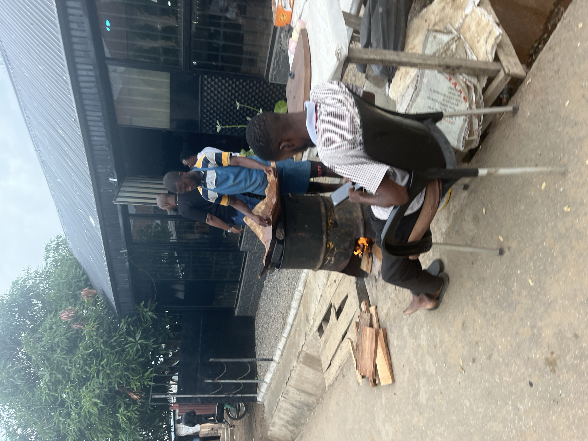 The image depicts an outdoor scene in Abuja, Nigeria, where a man wearing a striped t-shirt and dark pants is seated on a black plastic chair, holding a smartphone. He is positioned in the foreground, with his back partially turned to the camera. In the mid-ground, another man, wearing a blue apron over a t-shirt, is actively engaged in cooking over a makeshift grill fashioned from a metal drum. Flames are visible beneath the drum, indicating an active fire. Several pieces of wood are stacked on the ground near the cooking area, likely intended as fuel. Behind the cooking station, a building with a corrugated metal roof and glass windows is visible. The environment appears to be a street or roadside area, with a concrete gutter and pavement. Other individuals are present in the background, but their details are less discernible. The overall scene suggests a casual outdoor cooking or food preparation activity.