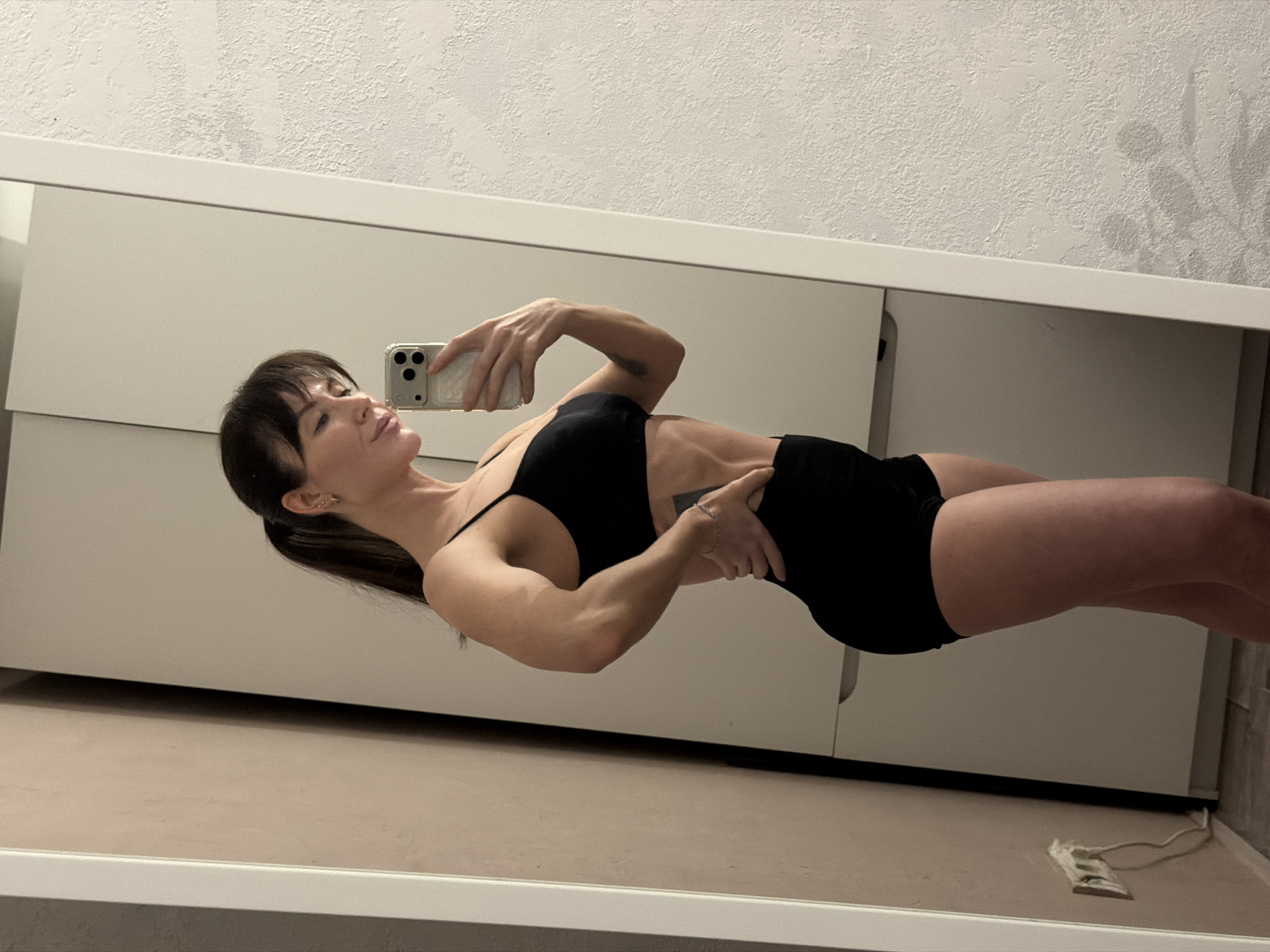 The image, captured in Perm, Russia, presents a mirror selfie of a woman taken indoors. The photograph is rotated 90 degrees clockwise, showing the woman's reflection in a horizontal orientation across the frame.
The woman, with dark hair styled with bangs and pulled back into a ponytail, is wearing a black sports bra or crop top and black shorts. Her physique appears toned and muscular, particularly visible in her arms and abdomen. She holds a light-colored smartphone, featuring multiple camera lenses, with her left hand, pointing it towards her reflection to capture the image. Her right hand is placed on her hip. Her facial expression is neutral, with a slight upward gaze and a subtle smile.
The background, visible in the mirror, consists of a white, textured wall above a white wardrobe or dresser unit that has prominent horizontal design lines. A light-colored, carpeted floor is also reflected at the bottom of the mirror. Faint shadows resembling foliage are cast on the textured wall in the upper right portion of the image. On the floor, near the bottom right, a white power cord or electronic device is visible. The setting appears to be an indoor room, likely illuminated by artificial light.