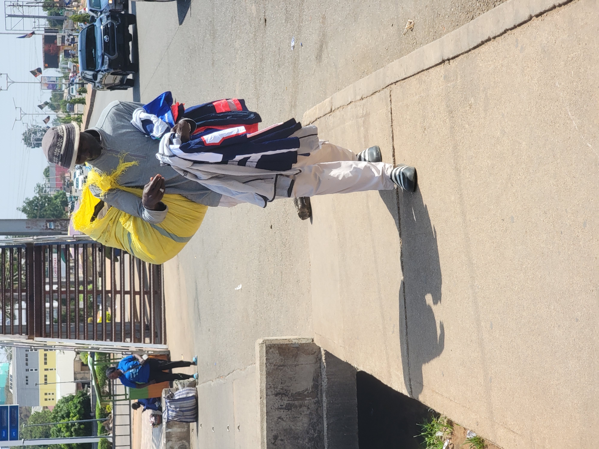 A man walks along a concrete sidewalk in Jos, Nigeria, carrying a large yellow bag and a stack of folded clothing. He is wearing a gray hat, long-sleeved gray shirt, and tan pants with black and white striped shoes. The street and sidewalk are concrete. In the background is a gate, other people, cars, and buildings in the city.