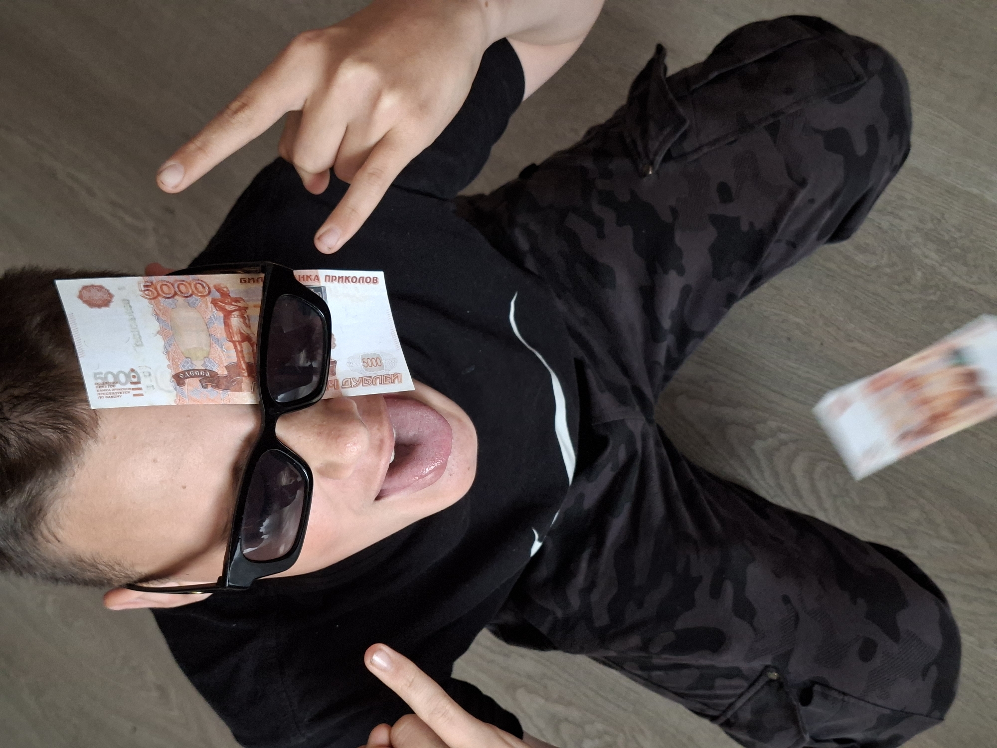 A young individual wearing black clothing and patterned trousers is lying on a wooden floor. They have a 5000 Russian Ruble banknote placed on their forehead and are wearing black sunglasses. Their tongue is sticking out. Both hands are raised, with the index and middle fingers extended, in what might be an obscene gesture. Another 5000 Russian Ruble banknote is out of focus to the right. The location is Unknown, Unknown.
