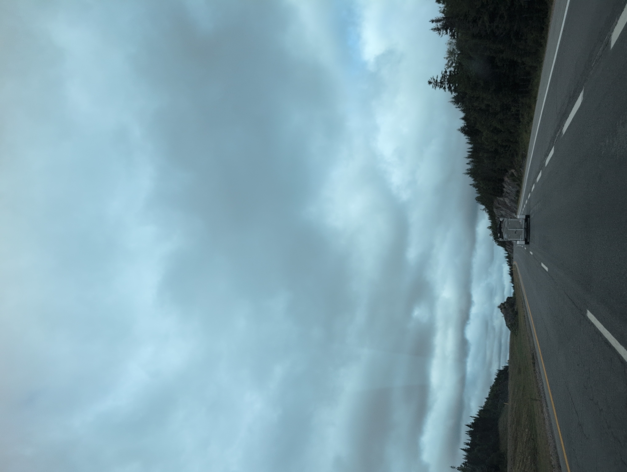 This image shows a view from a vehicle on a highway in Lepreau Parish, Canada, under a cloudy sky. The highway is paved with asphalt and has white lane markings. On the left side of the road, there is a grassy embankment with some trees and rocks. On the right side of the road, there is a dense forest. A silver trailer is attached to a vehicle in the distance, traveling in the same direction. The overall atmosphere is overcast and somber.