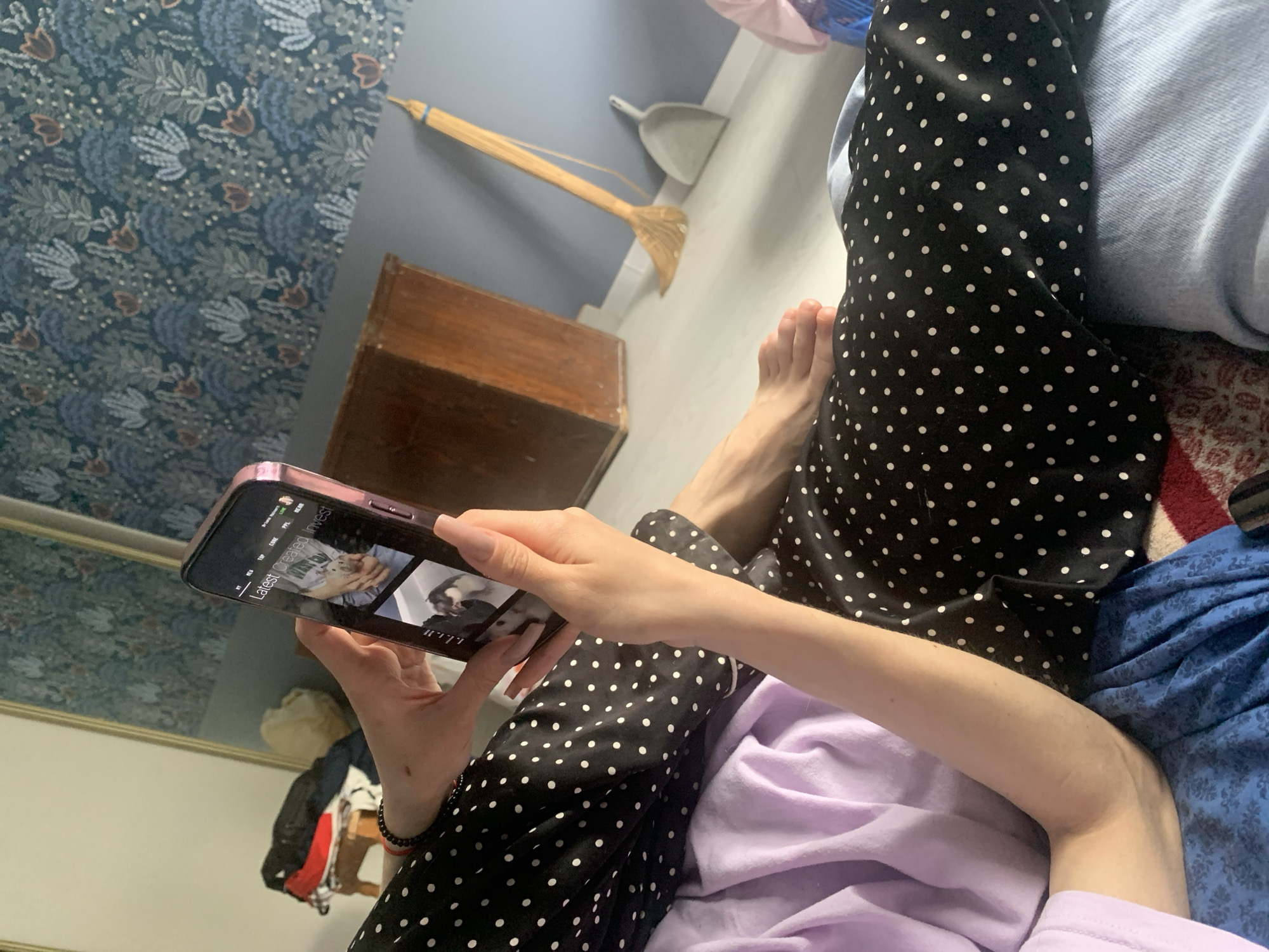 A person is holding a cell phone in their hands, viewing content titled "Latest created Invest." The person is wearing black and white polka-dot pants and a lilac-colored top. Their bare foot is visible. The scene also includes a wooden box, a broom, and a dustpan against a wall with blue patterned wallpaper. A mirror and clothes hanging are visible at the edge of the frame. There is no evidence to confirm this location is Stavropol, Russia.