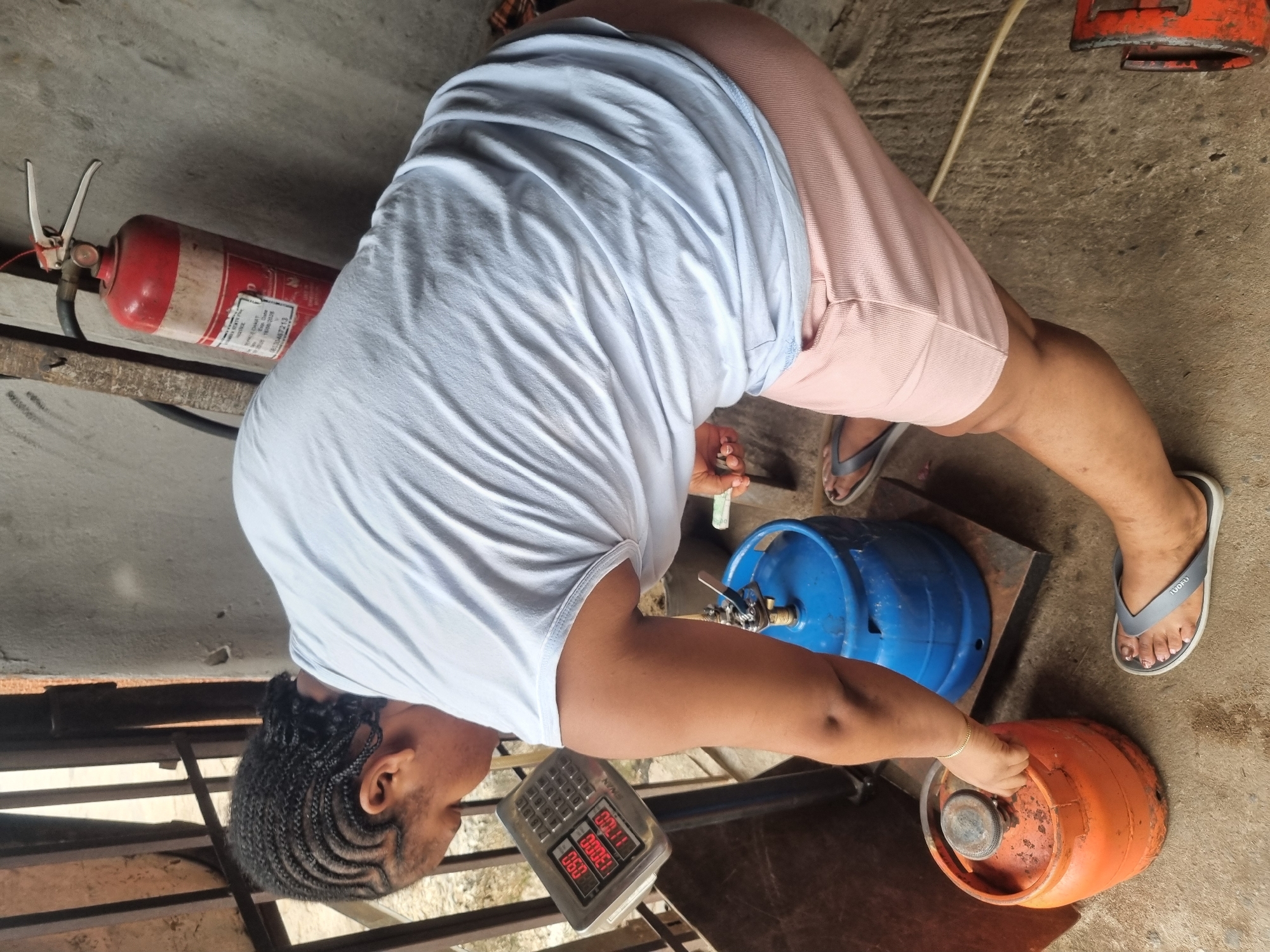 A person in a white t-shirt and pink shorts, wearing flip-flops, is transferring gas from an orange gas cylinder to a blue gas cylinder. The process is being monitored by a digital scale. A red fire extinguisher is visible in the background. The location is Onitsha, Nigeria.