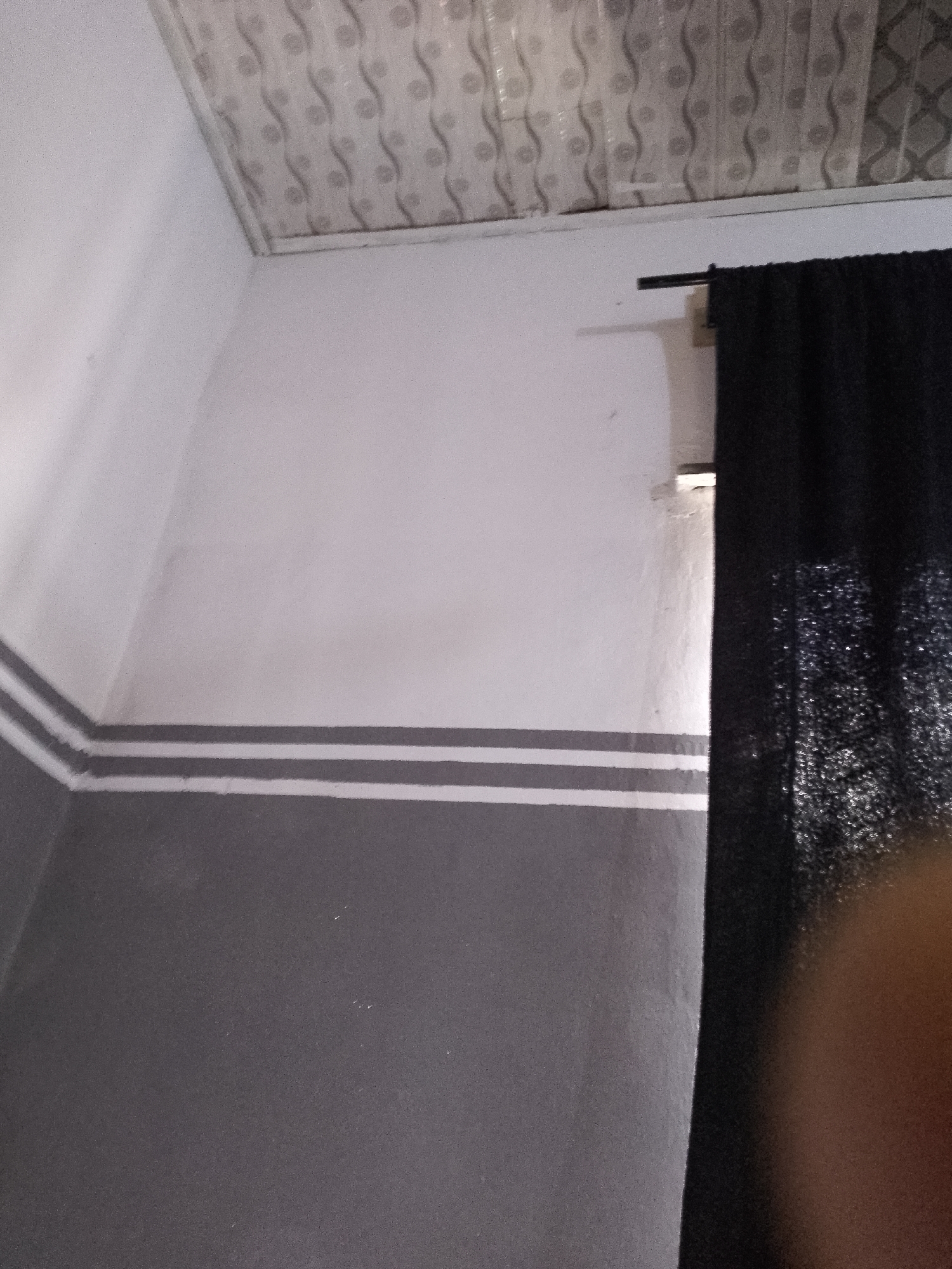 The image shows a corner of a room with a ceiling and two walls. The ceiling is white and has a patterned design. The walls are painted white and gray. There are three horizontal gray stripes on the white wall. A black curtain hangs on the right side of the image. The overall scene appears to be an indoor environment in an unknown location.