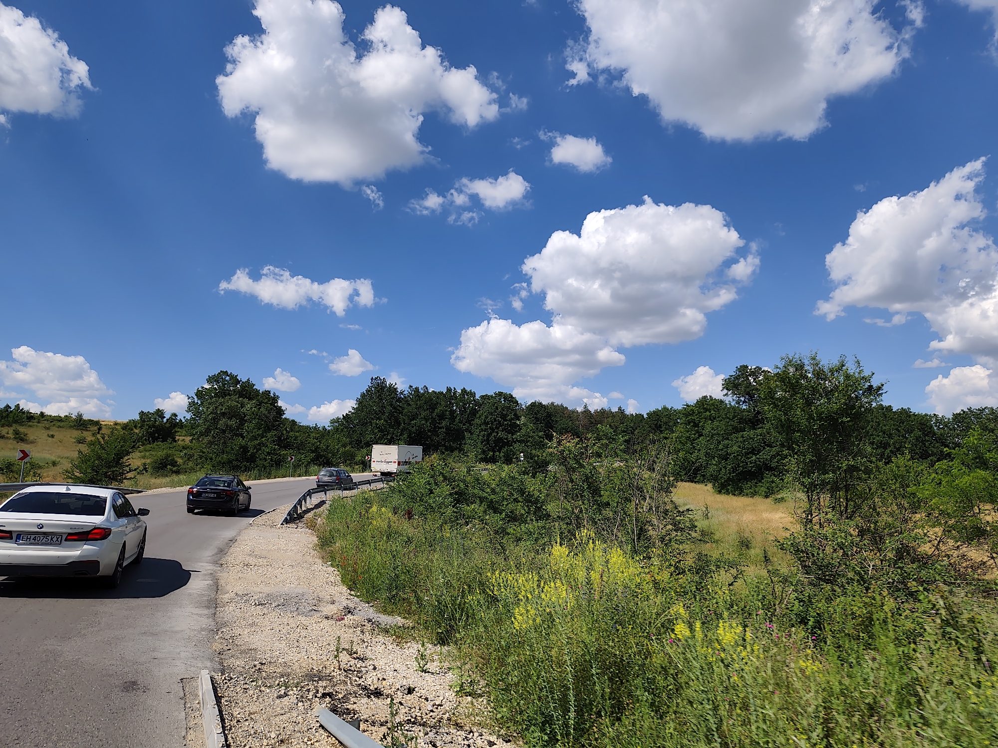 A daytime, eye-level shot captures a section of a paved road curving to the right, bordered by a gravel shoulder and dense green vegetation on the right. In the foreground, a white BMW sedan with Bulgarian license plate "EH 4075 KX" is driving away from the camera, its right turn signal illuminated. Further down the road, a dark sedan and a silver hatchback are also visible, along with a white truck. The scene is set against a backdrop of lush green trees and rolling hills under a bright blue sky filled with fluffy white clouds. The location is Aglen, Bulgaria.