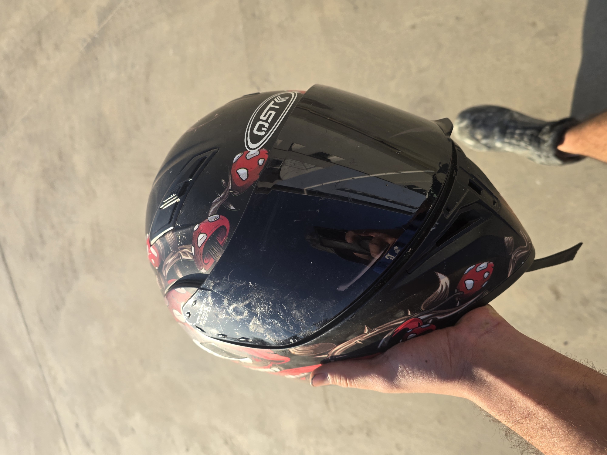 A person's hand holds a black full-face motorcycle helmet adorned with a red and white mushroom graphic. The helmet's visor is down and reflects the surrounding environment, including what appears to be a phone camera being used to take the picture. The helmet is decorated with what looks like a red mushroom with white spots, along with some foliage. The helmet also features a logo that reads "AST". The person is wearing a grey, skull-patterned clog. The background is a concrete surface, possibly a street or sidewalk, under bright sunlight, casting a shadow from the helmet. The scene is set in Yambol, Bulgaria.