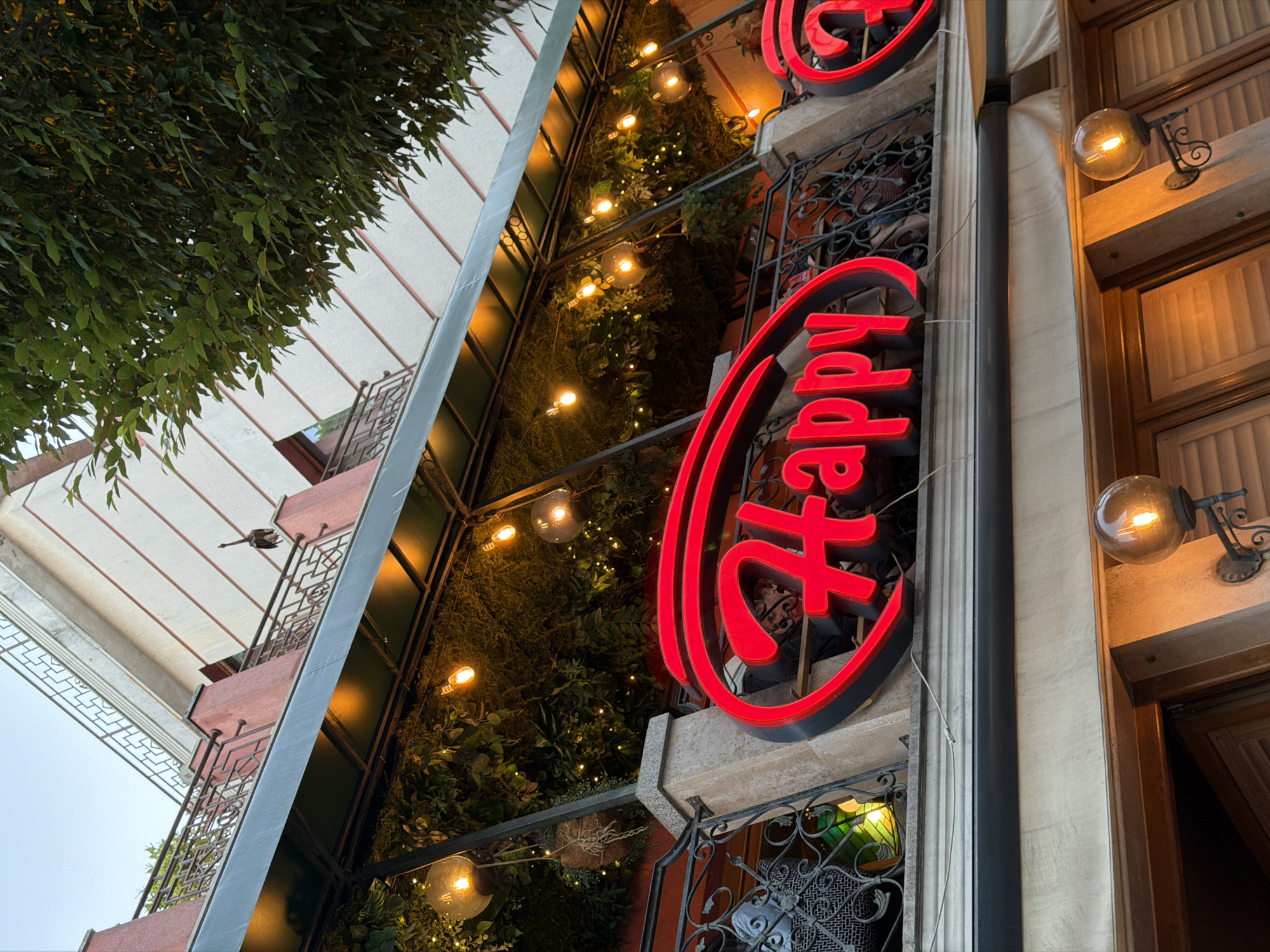 The image captures the exterior of a restaurant with a prominent red neon sign spelling out "Happy". The sign is illuminated, as are several globe-shaped light fixtures hanging from the ceiling of the covered outdoor seating area. Lush greenery, possibly plants or ivy, covers the facade behind the sign and the railing of the balcony above. A metal railing with decorative patterns runs along the balcony. The building itself appears to be several stories high, with a light-colored facade and red accents. A tree with dark green leaves is visible in the upper right corner of the frame, obscuring part of the building. The overall impression is of a vibrant, inviting establishment, likely a restaurant or cafe, located outdoors in a city setting. The specific location in Burgas, Bulgaria, is not discernable from the image itself.