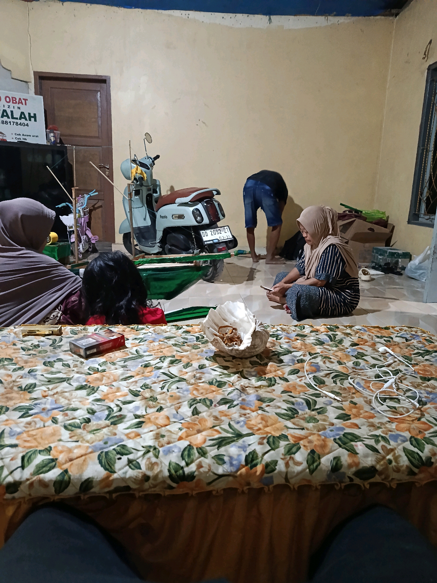 The image depicts an indoor scene, likely a living room or bedroom, in Bulukumba, Indonesia. In the foreground, a person's legs are visible, clad in dark blue jeans, suggesting they are the photographer or an observer. A mattress with a floral pattern covers the majority of the visible floor space. On the mattress, a pack of cigarettes and a tangled set of white earbuds are present. To the left, a woman wearing a grey hijab is partially visible. In the middle ground, a woman wearing a beige hijab and a striped shirt is seated on the floor, looking at her phone. Behind her, a light blue scooter is parked. To the left of the scooter, a child's bicycle with blue handlebars is visible. Further in the background, a man wearing dark shorts and a black t-shirt is bending over. A door is visible on the left wall, and a window with bars is on the right. The walls are painted a light beige color. The overall impression is of a casual, domestic gathering.