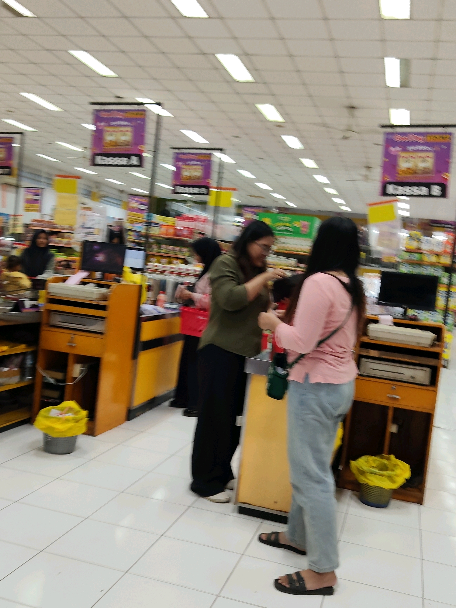 This is a medium-angle, indoor shot of a checkout area in a supermarket in Purwakarta, Indonesia. The scene is brightly lit by fluorescent ceiling lights. In the foreground, two women are interacting at a checkout counter. The woman on the right wears a pink long-sleeved shirt and light blue jeans, with a green crossbody bag. She holds a red shopping basket. The woman on the left wears a dark green long-sleeved shirt and black pants. She appears to be handing something to the other woman.
The checkout counter is made of orange wood with black accents. It has a cash register, a scanner, and shelves. To the left of the counter is a tall display shelf with various products. To the right of the counter, there is another checkout station with a computer monitor and a scanner. In the background, shelves stocked with products are visible, along with overhead signs indicating checkout lanes, one reading "Kassa A." A bright yellow trash bin with a yellow bag is positioned on the floor near the counter on the right. The floor is tiled in white. The overall atmosphere suggests a typical busy shopping environment.