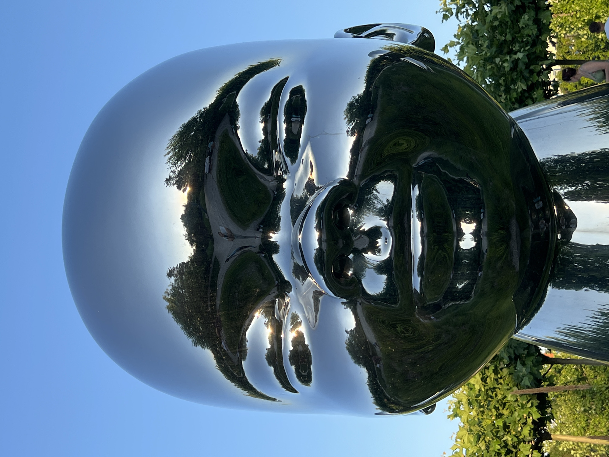 The image shows a large, highly polished, reflective sculpture of a human head, likely an artistic installation. The sculpture is outdoors, with a clear blue sky dominating the upper left portion of the frame. The surface of the sculpture acts as a mirror, reflecting the surrounding environment.
In the reflections, we can see trees with lush green foliage, and a paved pathway. Sunlight glints off the pathway and illuminates parts of the trees, suggesting it is daytime, possibly late afternoon given the angle of the sun. The reflections also capture distorted images of people, one of whom is visible on the right side of the frame, bending down. The sculpture itself is smooth and curved, with features like eyes, a nose, and lips subtly suggested by its form and the distortions in the reflection.
The overall scene is one of an outdoor public space, likely a park or a sculpture garden. The weather appears to be pleasant and sunny. The location is identified as Krasnodar, Russia. There is no visible text in the image. The mood is serene and contemplative, due to the still reflection of nature and the sky on the imposing sculpture.