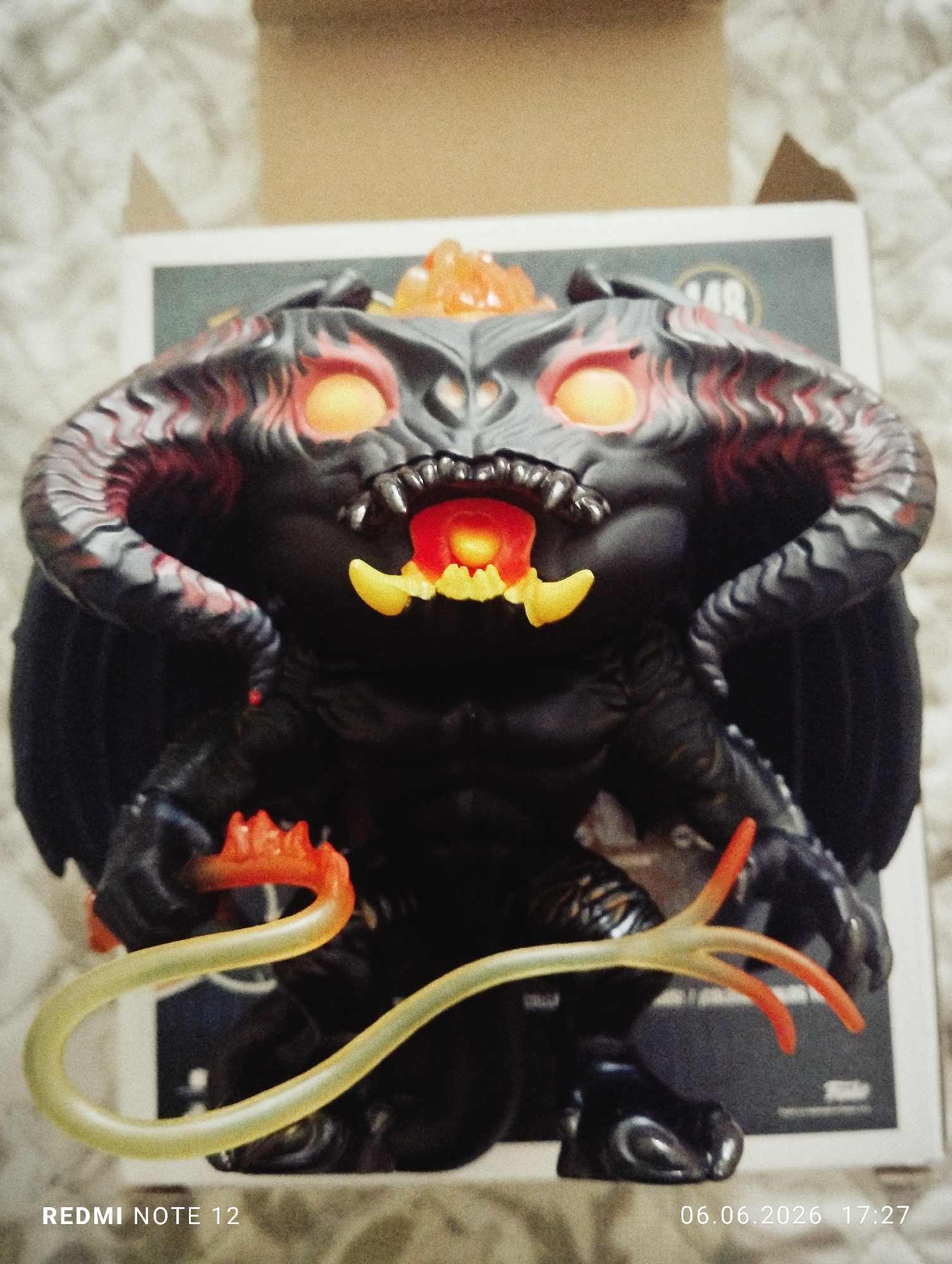 A forensic analysis of the image reveals a detailed close-up of a Funko Pop! vinyl figure, specifically a depiction of the Balrog from The Lord of the Rings, positioned indoors in the city of Burgas, Bulgaria. The figure is predominantly black, featuring large, textured horns, and prominent, rigid wings extending from its back. Its face presents large, glowing orange eyes and an open mouth with visible red and yellow fiery internal elements, along with sharp, yellow fangs. An orange, flame-like element emanates from the top of its head. The figure's right hand grasps a translucent, glowing yellow and orange whip, which extends across the lower left portion of the frame. The Balrog figure stands on a light-colored, subtly textured surface, likely a fabric, with its white and grey product packaging box visible directly behind it. The box displays the number '148' and a partial 'FUNKO' logo. In the upper background, a plain, light brown surface is discernible. The image was captured by a 'REDMI NOTE 12' device on '06.06.2026' at '17:27'.