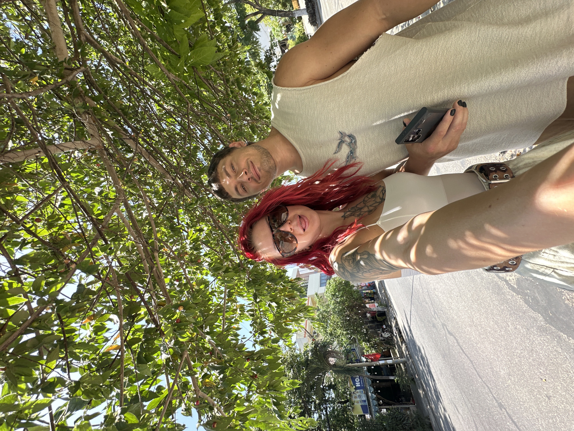 A low-angle, selfie-style shot captures two individuals standing on a paved road in Đà Nẵng, Vietnam, beneath the dappled shade of a leafy tree. The woman on the left, with vibrant red hair and sunglasses, is positioned closest to the camera, her left arm extended to hold the phone. She wears a white top and a belt with large circular buckles. Tattoos are visible on her left arm and chest. To her right, a man with short brown hair and a slight smile looks towards the camera. He is wearing a sleeveless beige shirt with a graphic print. He holds a dark-colored smartphone in his right hand, which is placed against his torso. In the background, a street lined with trees and buildings stretches out, with a sign for "HOA" visible on one of the establishments. The overall lighting suggests a bright, sunny day.