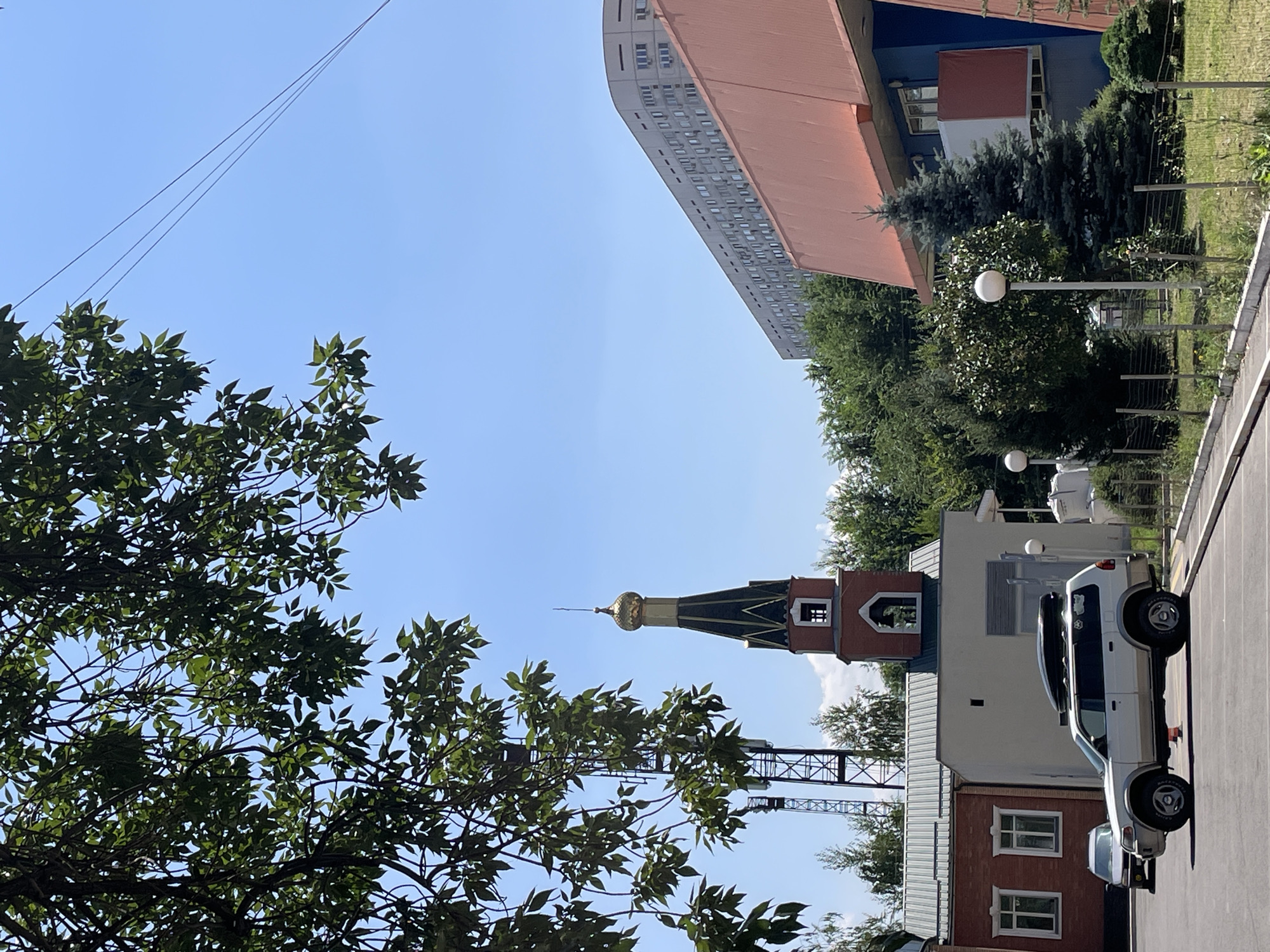 In Almaty, Kazakhstan, on a bright, sunny day, a scene unfolds with a mix of architectural styles and natural elements. In the foreground, a light-colored SUV with a roof-mounted cargo box is parked on an asphalt surface. To the left of the SUV, a smaller, dark-colored car is also visible. Behind them, a low-rise brick building with white-framed windows stands next to a lighter-colored structure.
Dominating the mid-ground is a bell tower with a red brick base and a green, octagonal roof topped with a golden dome, indicative of Orthodox Christian architecture. Adjacent to the bell tower, a tall, lattice-style tower, likely a telecommunications mast, rises against the clear blue sky.
Further in the background, a large, multi-story building with a curved facade and numerous windows dominates the skyline. In front of this building, and also to the right of the scene, are lush green trees, suggesting a well-vegetated area. A modern building with a vibrant orange roof and blue accents is situated on the right side, partially obscuring the view of the taller building. The ground in the foreground features patchy grass and a metal fence. Overhead, tree branches with green leaves frame the top left portion of the image, and thin utility lines are visible against the sky.