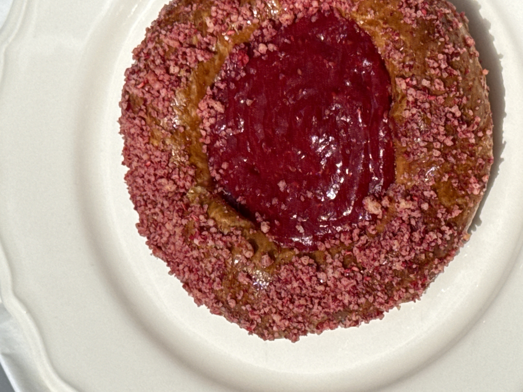 A close-up, overhead shot shows a round pastry with a red berry filling and a crust coated in pink, granulated pieces, resting on a white ceramic plate. The pastry is centrally located on the plate, which is set against a white, slightly textured surface. The lighting casts a soft shadow to the right of the pastry, indicating a light source from the upper left. The pastry itself is golden brown with a slightly glossy texture, and the filling appears smooth and viscous. The pink coating is unevenly distributed around the edge of the pastry, creating a textured border. No people or other objects are visible in the image. The location is not specified, and Vracar Urban Municipality, Serbia is not depicted.