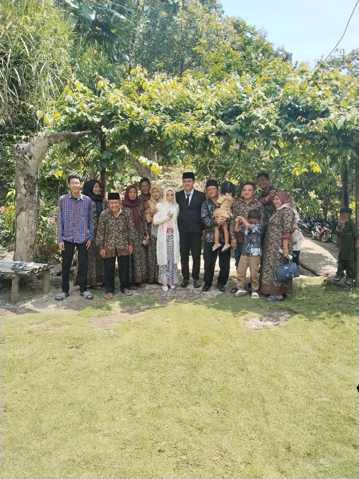 A group of fourteen people is posing for a photo outdoors in a grassy area under a tree with dense foliage. The location is Unknown, Unknown. The subjects include men, women, and children dressed in a mix of casual and formal attire. The central figures appear to be a bride and groom, the bride wearing a white headscarf and dress with a patterned overlay, and the groom in a black suit and head covering. Some individuals are wearing traditional batik patterns. A child is being held by an adult. In the background, there are parked motorcycles and a glimpse of buildings. A young boy is standing apart from the main group. A bamboo bench is visible in the left background. The overall lighting suggests a daytime setting.