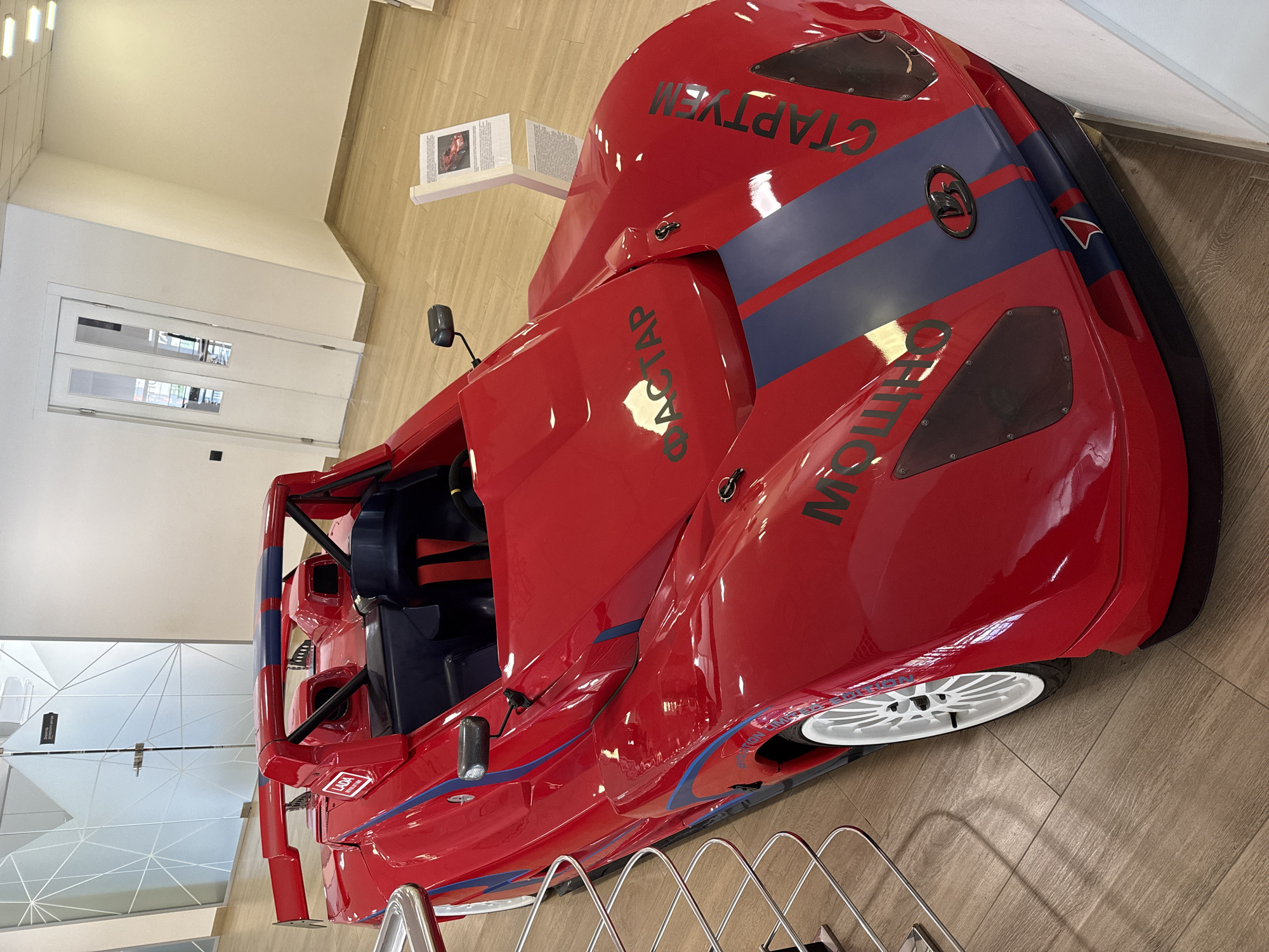 The image shows a red racing car on display in what appears to be an indoor exhibition space in the city of Unknown, Unknown. The car has blue stripes and lettering in Cyrillic. Visible parts of the interior include blue seats and a roll cage. The floor is a light brown, wood-look material. In the background, there is a white wall, a door, and a display stand with informational text. A metal barrier surrounds the car.