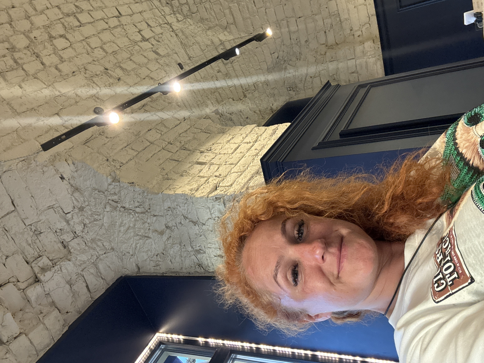 A woman with vibrant red, curly hair is captured in a selfie, her face turned slightly towards the camera and a subtle smile gracing her lips. She wears a white t-shirt with a graphic print. The background reveals an interior space with a rustic exposed brick ceiling arching overhead. Dark blue walls frame the scene, and a black cabinet stands to the right. Track lighting with spotlights illuminates the brickwork. The image's composition, with the woman's face in the foreground and the architectural details behind her, suggests she is indoors, possibly in a restaurant or a historical building.