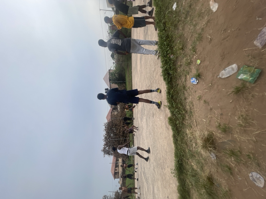 The image depicts an outdoor scene in Umunekwu, Nigeria, on an overcast day. In the foreground, a strip of unkempt dirt and sparse green grass is visible, littered with several plastic bottles and other discarded packaging. This area slopes upward to a worn, light-colored concrete or asphalt court occupying the midground.
On the court, approximately 15-20 young adult males are engaged in or observing an activity, likely a sport. Many individuals are dressed in athletic wear, including t-shirts, tank tops, and shorts, with some shirtless. Footwear primarily consists of sneakers.
Prominently in the center foreground, with his back to the viewer, stands a male wearing a dark blue top, dark shorts, and athletic shoes with patterned socks. To his left, another male in a white tank top and light grey shorts is in motion, facing right. On the right side of the frame, three males are standing, observing the court: one in a grey t-shirt with partially visible text, light grey sweatpants, and hands on hips; next to him, a male in a yellow t-shirt and dark shorts; and further right, a male in a dark top and shorts. Further back on the court, multiple individuals are grouped in various postures suggesting active play or gathering.
The background features dense, dark green trees and bushes that partially obscure several residential-style buildings with reddish-brown roofs, indicating a suburban environment. A utility pole with wires is visible on the far right, and a dark water tank is partially visible near one of the background buildings. The sky is uniformly light grey, suggesting an overcast condition.