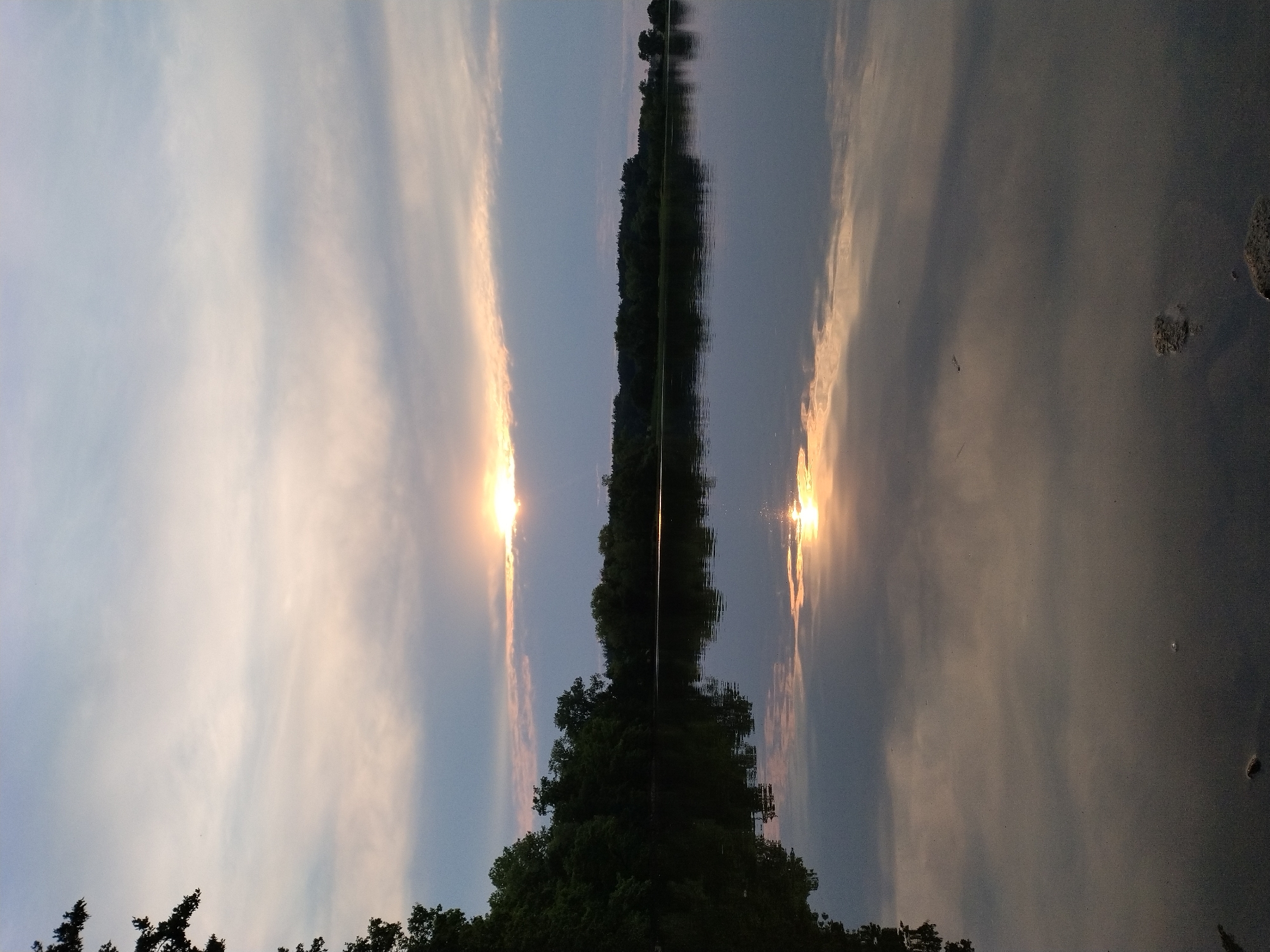 This is a vertical, wide-angle shot of a lake at dusk, likely taken in Kestřany, Czechia, as suggested by the provided context. The sun is setting behind a strip of clouds, casting a warm, golden glow on the sky. The water of the lake is calm, reflecting the sky and the tree line on the opposite shore. The foreground shows a few rocks and debris on the lakebed, partially submerged. The left side of the image is dominated by the dark silhouette of a tree and dense foliage. The overall mood is serene and peaceful, characteristic of a natural landscape at twilight. There are no people or animals visible in the frame.