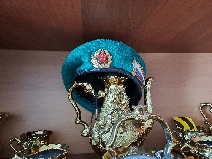 This image depicts an indoor scene in the city of Полянки, Russia, showcasing several decorative objects displayed on a wooden shelf. A teal-colored military beret is prominently featured, resting on a highly ornate, golden, metallic decorative container that resembles a teapot or trophy, adorned with intricate relief carvings. The beret displays a central emblem: a red star containing a gold hammer and sickle, surrounded by a gold wreath, consistent with Soviet-era military insignia. A small patch, resembling the Russian tricolor flag (white, blue, red), is attached to the right side of the beret. To the immediate left, a portion of a smaller, dark-colored decorative cup or vase with golden scalloped edges and floral patterns is visible. To the right, part of another silver-colored ornate object is present, with a yellow and black striped object partially discernible behind it. The objects are situated on a light brown wooden shelf, with a darker brown wooden surface visible above, suggesting a display cabinet or shelving unit. No people or active interactions are visible.