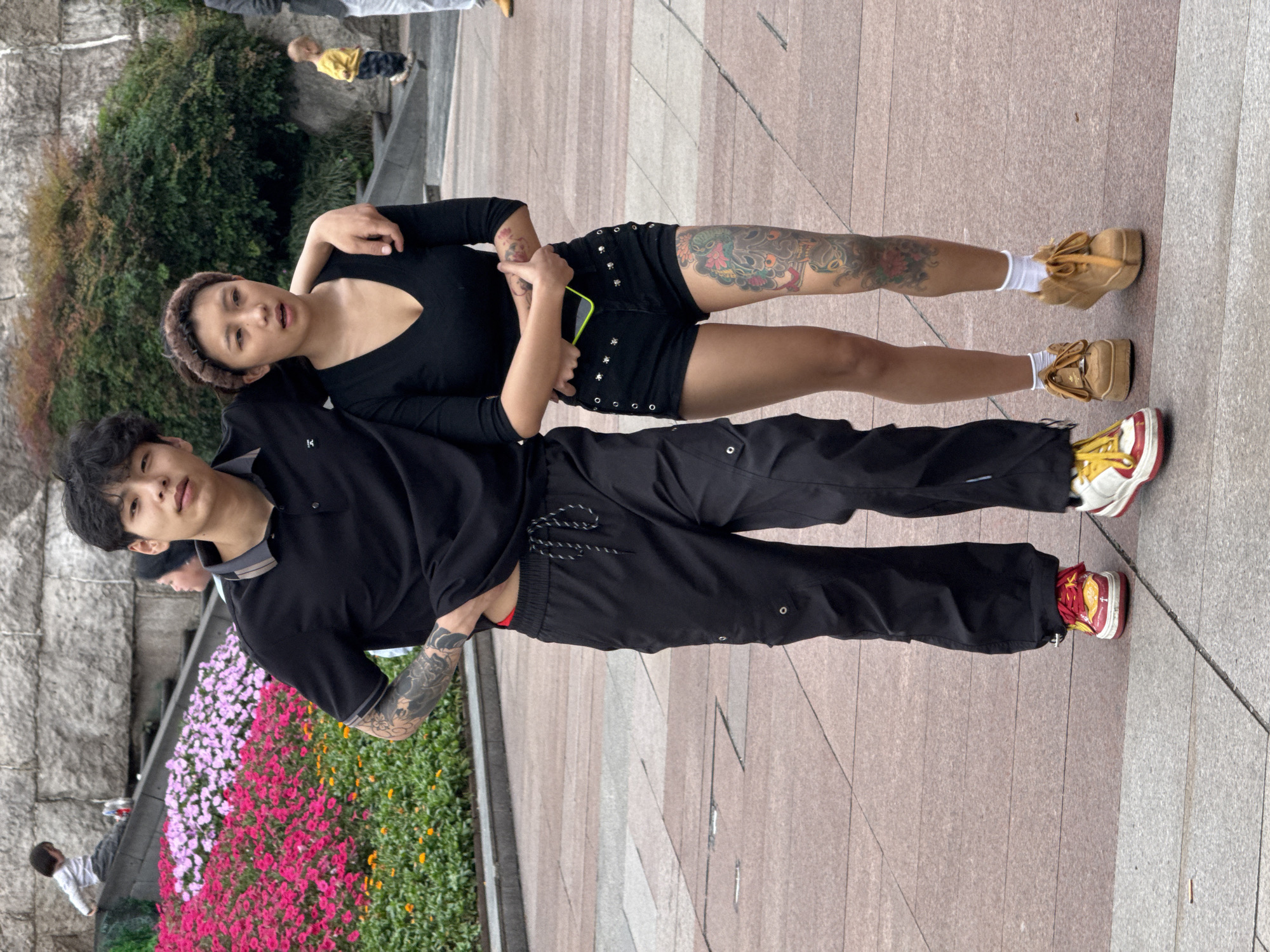 Two people are standing close to each other in an outdoor environment. A person wearing a black short-sleeved shirt and black pants with red and white sneakers is standing next to a person in a black long-sleeved shirt and black shorts with beige platform shoes. Both individuals have tattoos. The location appears to be a public area in Liangjiang, China, near flower beds and stone structures.