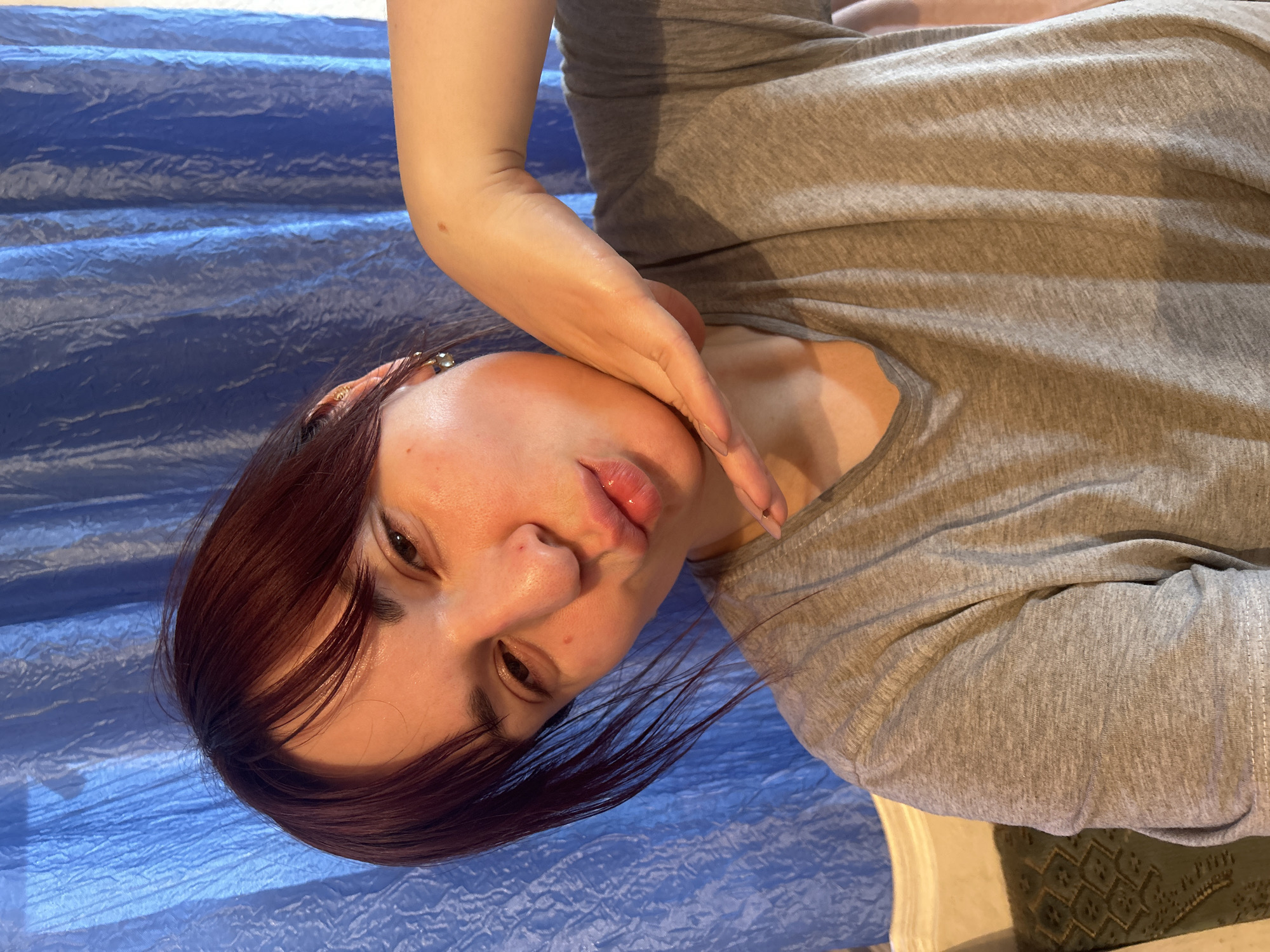 A close-up, portrait-style image features a person, likely female, with a pensive expression. The individual has medium-length, dark reddish-brown hair with bangs, styled to sweep across the forehead. One eye is partially visible, gazing towards the viewer, while the other is mostly obscured by hair and the angle of the head. The lips are slightly parted, appearing full and glossy. A hand, with manicured nails, is raised to the side of the face and neck, creating a posed or thoughtful gesture. The person is wearing a simple, light grey t-shirt or top made of a textured, possibly cotton, fabric.
The background predominantly features a deeply crinkled, textured blue surface, resembling a tarp or a rumpled sheet. In the lower right corner, a small section of a light-colored object, possibly furniture, and a dark, patterned textile (potentially upholstery or a cushion) are visible, suggesting an indoor setting. The lighting is soft and even, without harsh shadows, indicating either indirect natural light or artificial illumination. The mood conveyed is calm and introspective. There is no visible text in the image.