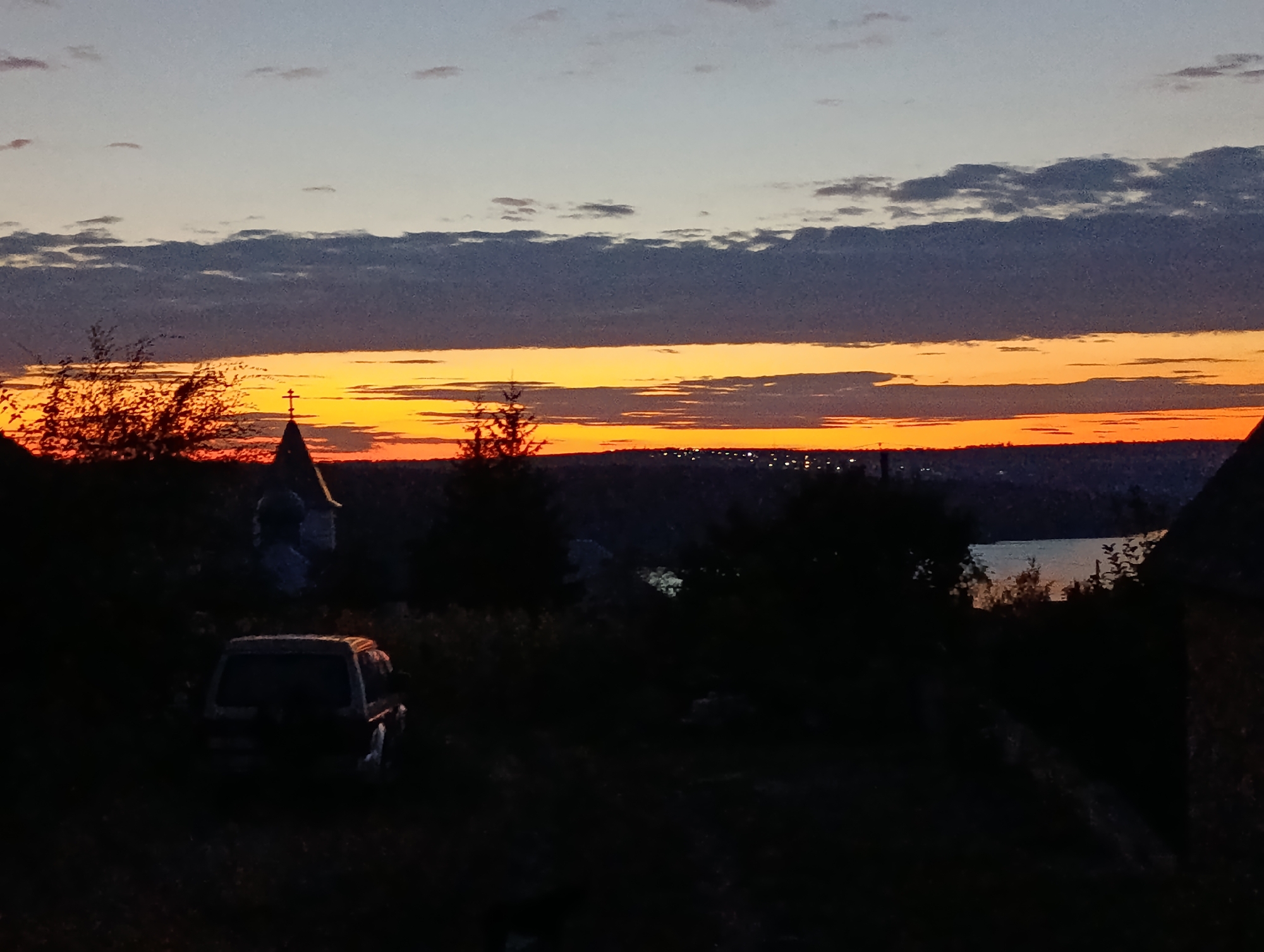 This is a photograph of a sunset over a landscape, likely in the city of Răscăieți, Moldova. The sky is a gradient of colors, with a pale blue at the top fading into shades of orange and red near the horizon. Dark, silhouetted trees and bushes are in the foreground, obscuring much of the ground. A dark-colored SUV is parked on the left side of the frame, partially visible in the shadows. In the mid-ground, a church spire with a cross can be seen against the bright sky, along with more trees. The horizon is dotted with lights, suggesting a settlement. To the right, the dark outline of a building is visible. The overall atmosphere is one of twilight and quietude.