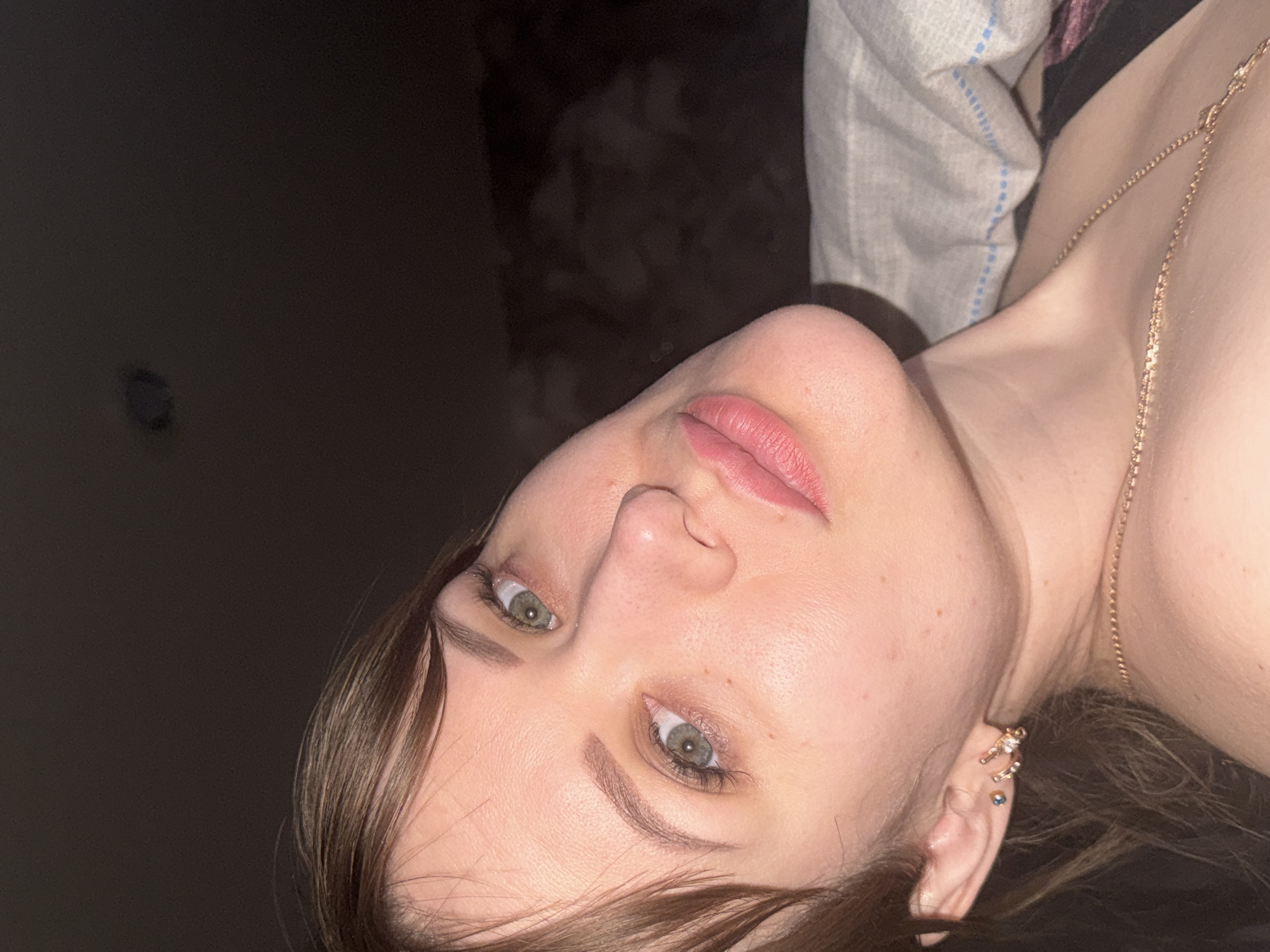 A close-up photograph depicts a woman from a low angle, focusing on her face and upper chest. Her head is tilted slightly to the right. She has light skin, brown hair with bangs, green eyes, and pink lips. A gold chain necklace is visible on her chest. She is wearing multiple earrings in her left ear. The background includes a dark wall and a light-colored pillow with blue stitching. There is no indication of the location being Chaikovsky, Russia.