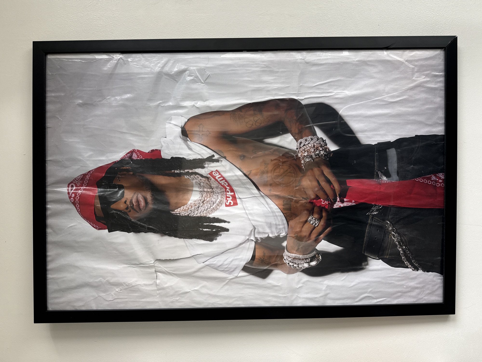 A vertical photograph captures a black-framed artwork displayed on a plain, light beige wall. The artwork is a full-body portrait of a male subject with long, dark dreadlocks, dark skin, and a light beard. He wears a red paisley bandana on his head, dark rectangular sunglasses, and a visible philtrum piercing. The subject is heavily adorned with multiple diamond-encrusted chains around his neck, numerous iced-out bracelets on both wrists, and several prominent rings on his fingers. He wears a white t-shirt with a red "Supreme" box logo, which is pulled up, exposing his abdomen and lower chest. Visible tattoos cover his arms, chest, and abdomen. His hands are positioned near his waist, appearing to adjust or hold a red bandana that is tucked into dark pants, which are accessorized with a silver chain. The background within the portrait is a deliberately creased, wrinkled white or light gray surface, mimicking a distressed street poster. Shadows are cast by the subject onto this background as part of the artwork's composition. The image provides no visual indicators to determine a specific location within New York City, United States of America.