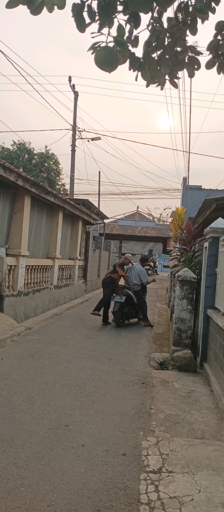 Two men are standing on a narrow asphalt road in what appears to be a residential area, possibly in Subang, Indonesia. The man on the left is wearing a patterned short-sleeved shirt and dark pants, with his left leg extended as if he is about to step off a motorcycle. The man on the right is wearing a light-colored short-sleeved shirt and dark pants, and is leaning over the motorcycle. The motorcycle is dark-colored with a license plate that reads "2230 YM". The road is flanked by buildings on both sides, one with a light-colored facade and a railing, and another with a darker color. Overhead, a complex network of electrical wires is visible against a pale sky, with the sun faintly visible through the haze. In the foreground, leaves from a tree with round fruit hang down, partially obscuring the view. The overall impression is that of a casual encounter or repair on a quiet street.