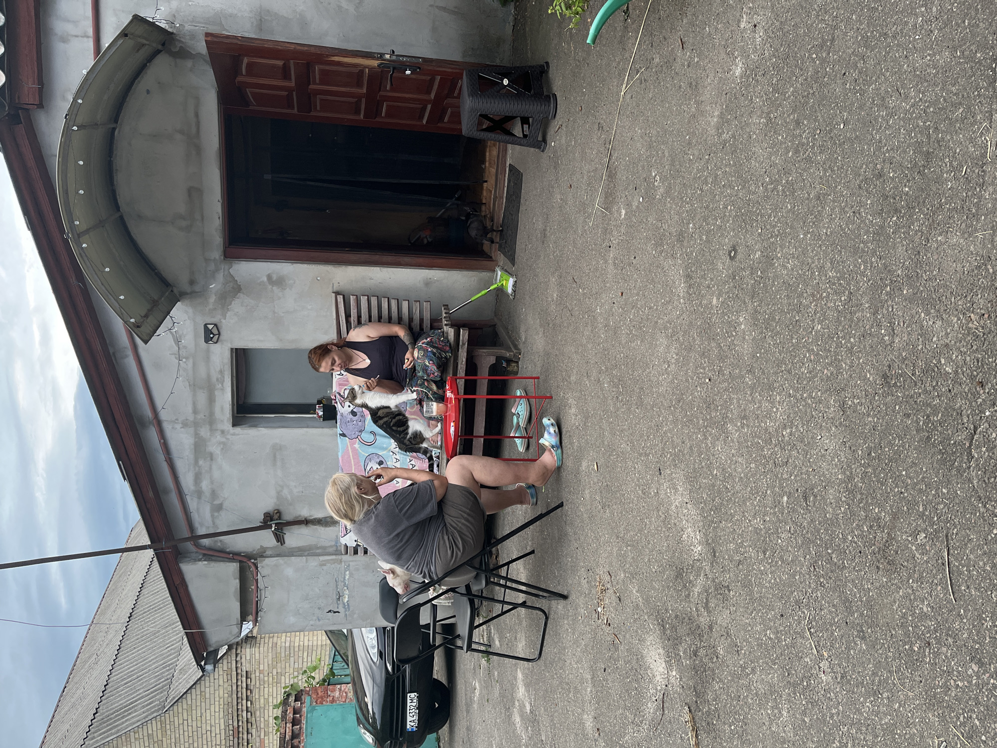 In Nemishaieve, Ukraine, an overcast day features an outdoor scene in front of a weathered concrete building with a wooden door and an arched awning adorned with string lights. Two women are seated outside, one in a gray t-shirt and shorts, the other in a black tank top. A cat sits on the lap of the woman in the black tank top, and another cat is visible on a chair nearby. A small red table with a drink on it separates them. To the right, a black stool and a mop with a green head rest on the pavement. A black car with a Ukrainian license plate is parked to the left of the building. The ground is paved with asphalt, and there is some debris scattered about.