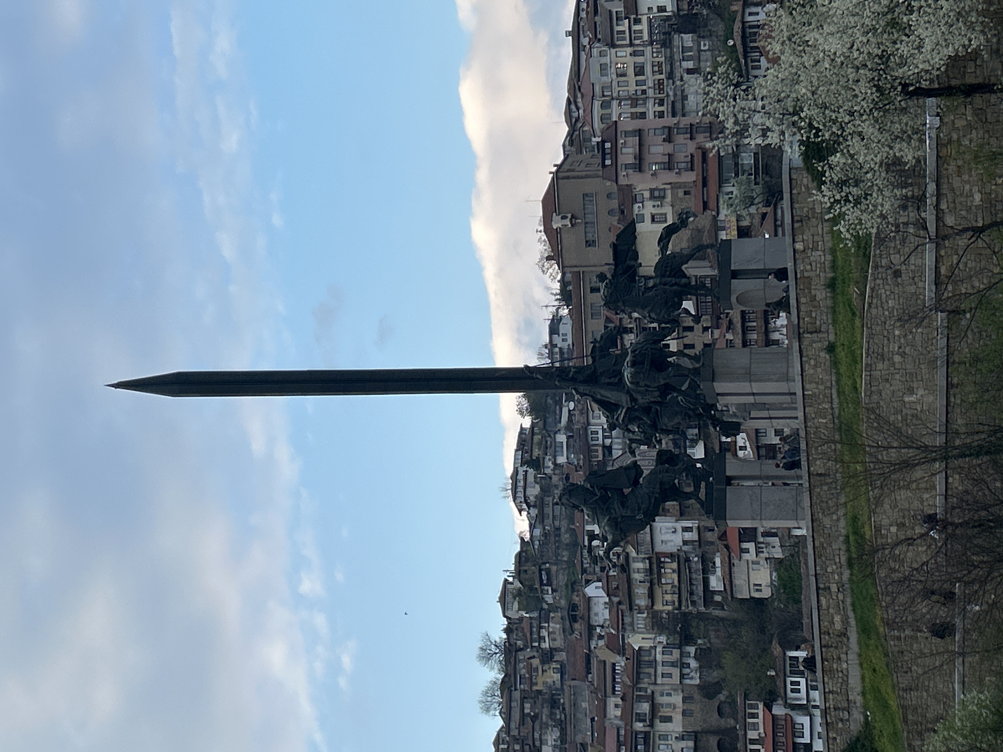 The image depicts the Asenevtsi Monument in Veliko Tarnovo, Bulgaria, against a backdrop of a hillside densely populated with buildings and a cloudy sky. The monument, featuring a large sword and equestrian figures, is prominently displayed in the foreground. A stone wall runs along the right side of the image, with a blossoming tree partially visible. The buildings on the hillside show a mix of architectural styles and colors.