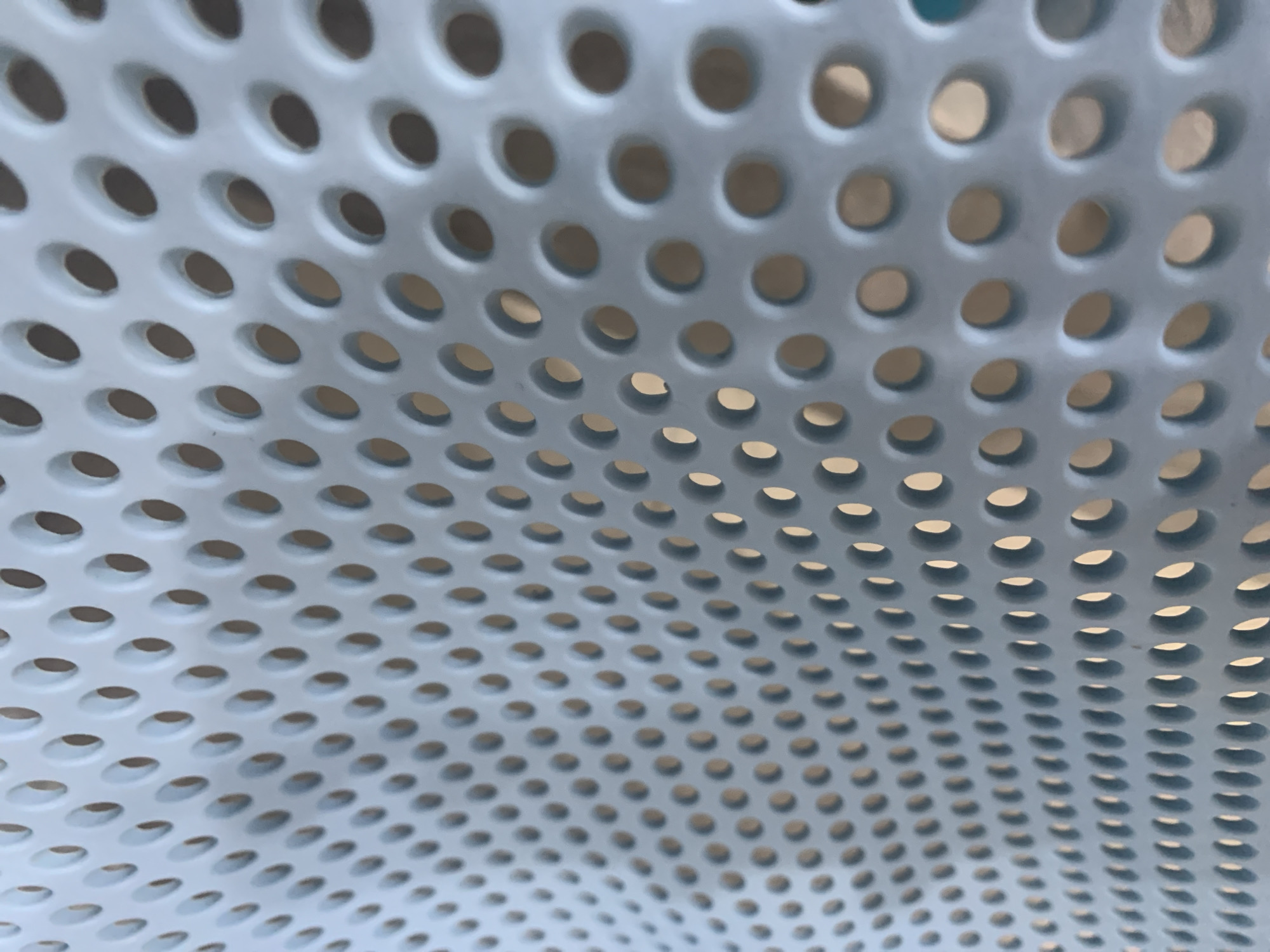 The image displays a close-up, abstract view of a light blue, perforated surface. The surface is curved, creating a sense of depth and perspective. The perforations are small, oval-shaped holes arranged in a regular grid pattern. Through the holes, a warmer-toned background is visible, suggesting a different material or space behind the perforated surface. The lighting casts subtle shadows around the edges of the holes, highlighting their three-dimensional quality. The overall impression is one of a textured, industrial, or architectural detail, possibly a part of furniture, a screen, or a decorative panel. No people, specific objects, or environmental details like time of day or weather are discernible. There is no visible text in the image. The provided location context of Stavropol, Russia, does not offer any direct visual cues within the image itself.