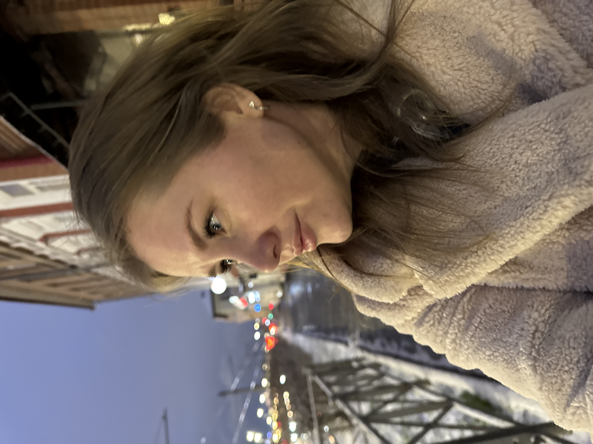 This media file features a portrait of a young woman taken outdoors at dusk or night in Perm, Russia. The woman, positioned prominently on the left and center of the frame, has long, wavy light brown hair. She possesses fair skin, light-colored eyes enhanced with visible eyeliner and mascara, and slightly parted lips, conveying a soft, contemplative expression as she looks downwards and to her left. She is dressed in a warm, fluffy coat or jacket of a light beige or cream color, and a small silver stud earring is visible in her right ear.
The background is softly blurred, creating a bokeh effect with numerous out-of-focus lights in vibrant red, white, blue, and yellow hues, suggesting an urban street scene with traffic and streetlights. Faint outlines of a road or street are visible, and white, textured areas indicate snow covering the ground, hinting at cold weather. Dark, linear structures, possibly a bridge railing or other urban infrastructure, are dimly discernible in the lower right background. The sky is a deep blue, characteristic of evening. The scene captures a serene moment within a bustling, cold urban environment. There is no visible text.
