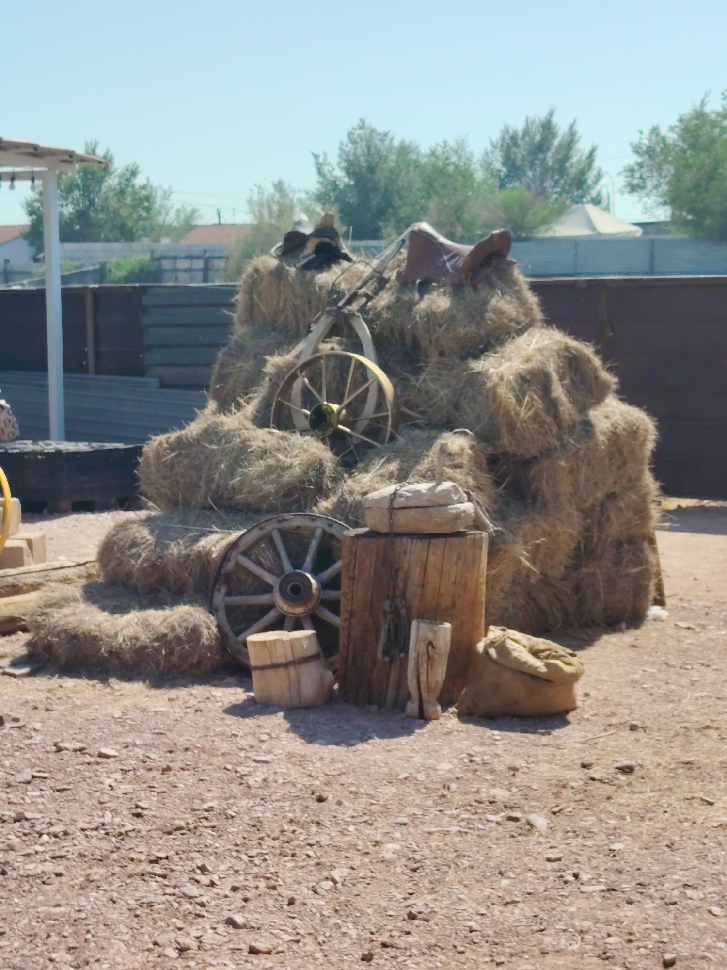 The image depicts a rustic scene with a large stack of hay bales. A vintage wagon wheel with spokes is positioned in front of the hay, and another wheel is embedded in the hay on the right side. A saddle rests on top of the hay stack. In the foreground, there is a wooden stump with some tools attached, and a burlap sack is placed on the ground beside it. The ground is covered with gravel and small stones. In the background, there is a wooden fence and some trees, with buildings and a light-colored tent visible further beyond. The sky is clear and blue. The overall impression is an outdoor setting that evokes a historical or rural ambiance. There are no people visible in the image. The location is identified as Toranghylyq, Kazakhstan.
