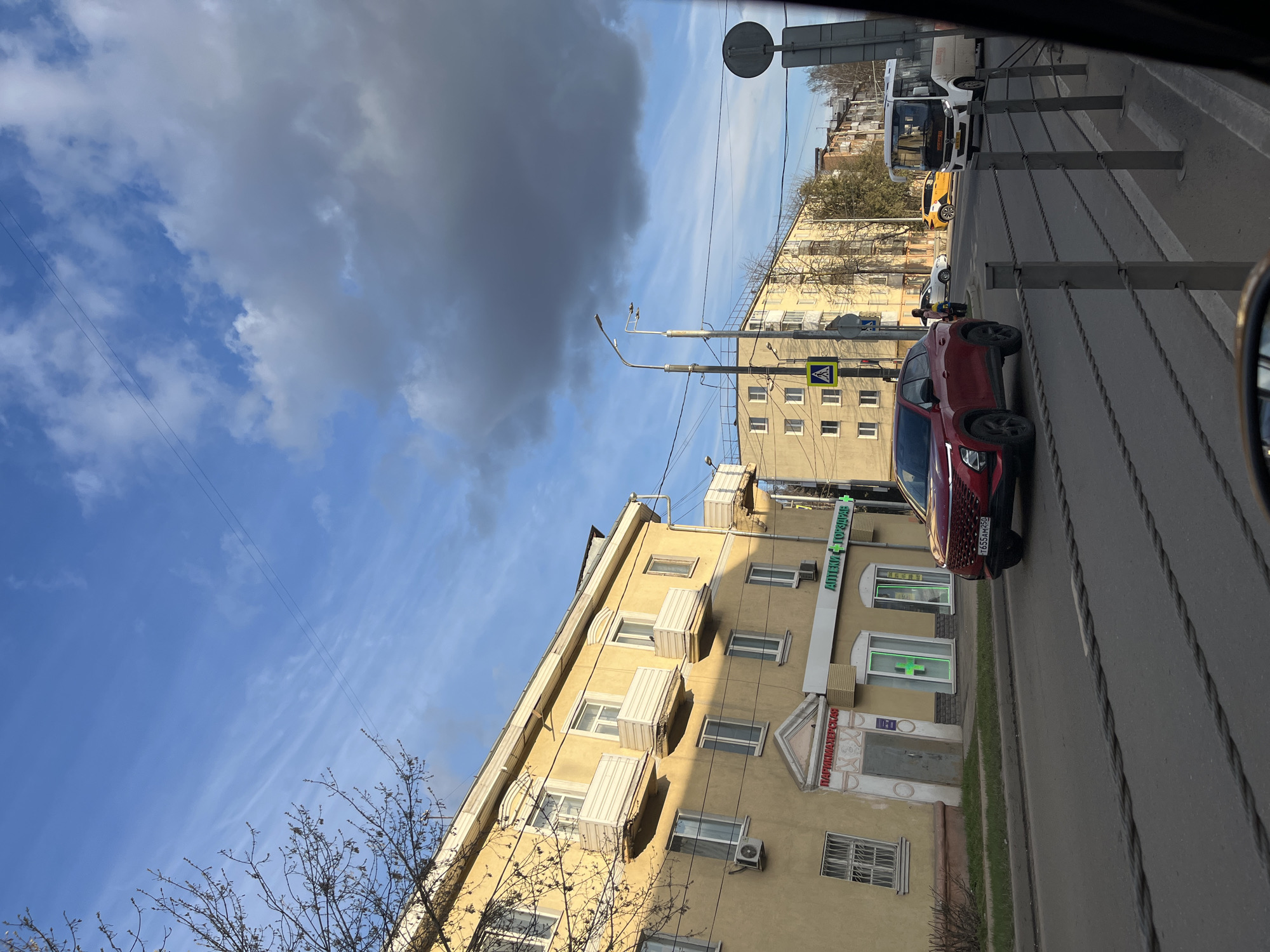 The image shows a street scene in Klin, Russia. A red car is parked along the right side of the road in front of a yellow building with a sign in Russian for a pharmacy ("АПТЕКА" in green). The building has multiple windows with what appear to be air conditioning units installed below them. Across the street, there are other buildings, as well as a yellow taxi and a white van. A pedestrian crossing sign is visible, and power lines stretch across the blue sky with scattered clouds. A metal barrier lines the sidewalk. The perspective suggests the photo was taken from inside a car.