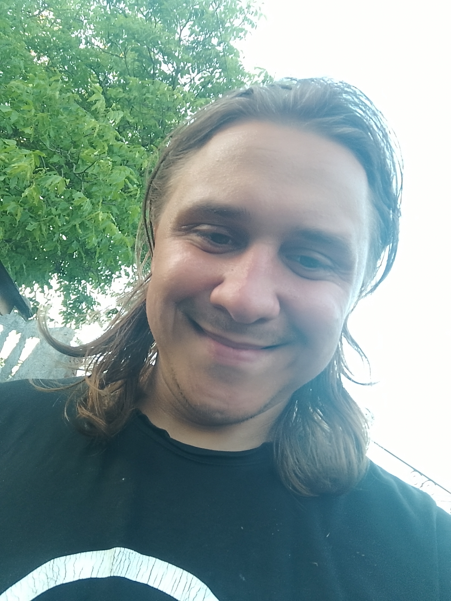 A close-up selfie shows a light-skinned man with long brown hair, a mustache, and a beard stubble. He is wearing a black t-shirt with a white circular graphic on the front. He has a relaxed smile. The background includes lush green foliage, suggesting an outdoor setting. A white sky is visible in the upper right portion of the frame. The man is looking directly at the camera. The image appears to have been taken during daylight. The specific location in the city of Dnipro, Ukraine is not discernible from the image itself.