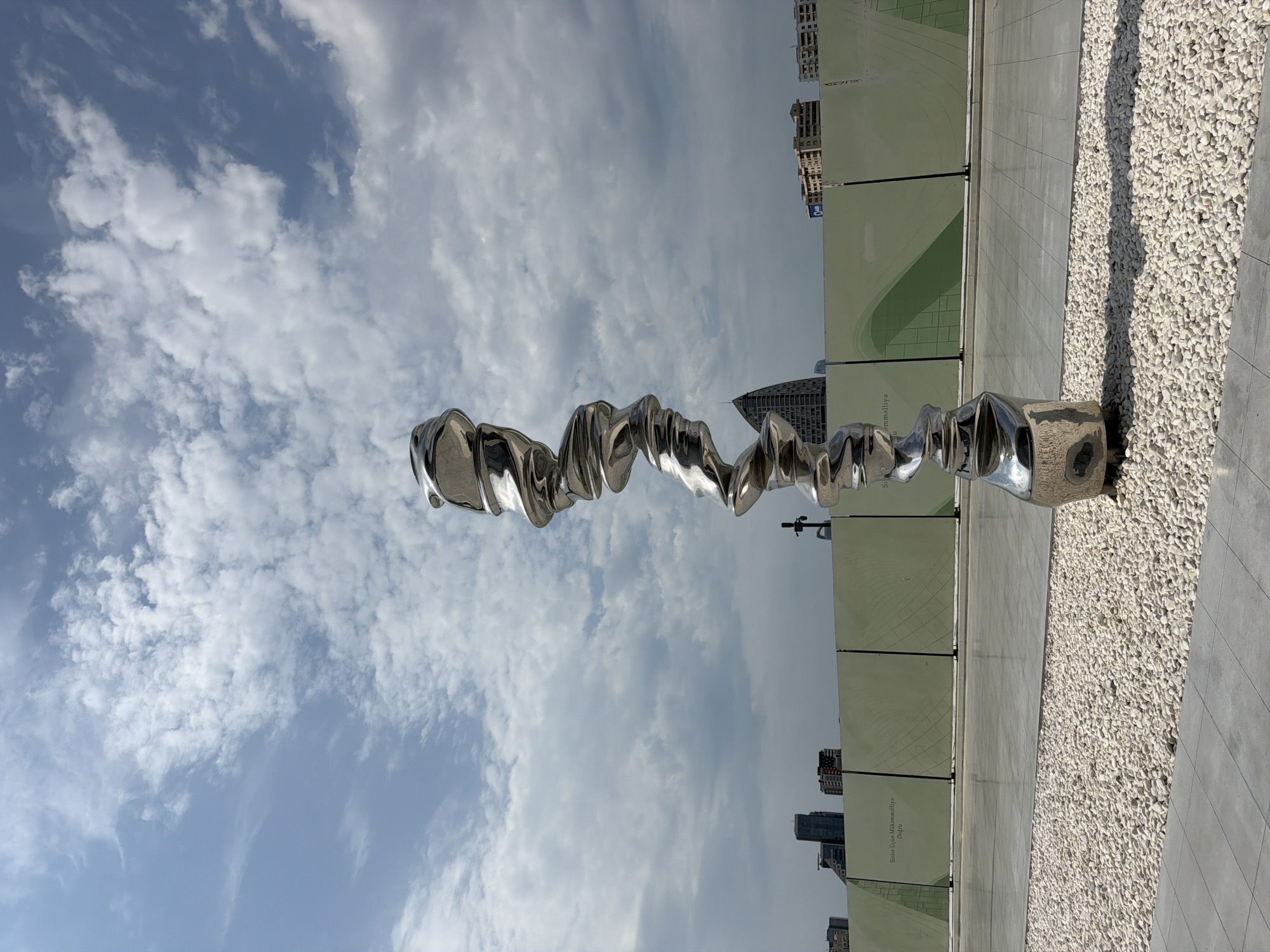A tall, abstract, chrome sculpture stands in the foreground against a backdrop of a cloudy blue sky, buildings, and a large green construction barrier. The sculpture is spiraled and twisted, resembling a distorted human form. It is positioned on a paved area next to a section of white gravel. In the mid-ground, a light green construction barrier is visible, with text in a foreign language and a green graphic design. Beyond the barrier, city buildings of various heights and architectural styles are silhouetted against the sky. The overall scene suggests an outdoor urban environment, possibly a public space undergoing development or renovation. The location is identified as Baku, Azerbaijan. There are no people or animals visible in the image. The lighting indicates it is daytime, with scattered clouds creating some shadows on the ground.