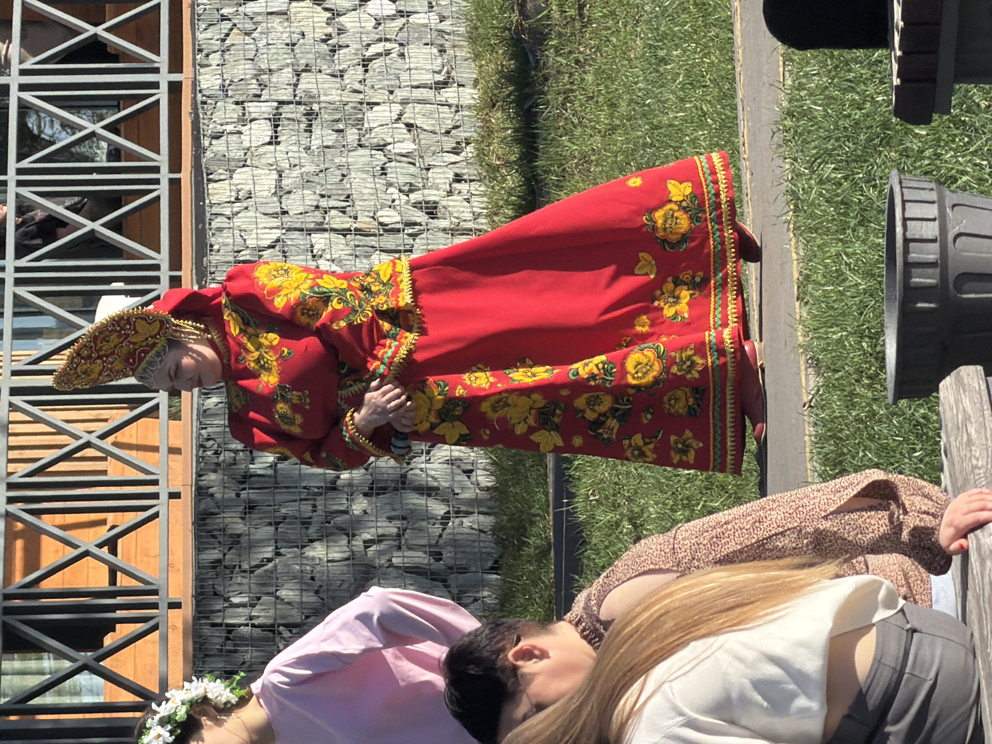 A woman in traditional Russian clothing stands outdoors in Votkinsk, Russia. The woman wears a long red dress adorned with yellow floral patterns, gold trim, and a matching headdress. Her hands are clasped in front of her. Behind her are a building with a metal framework and a wall of stacked stones. To the right of the woman is grass and a wooden bench. In the foreground are two women; one wears a pink top and a floral headpiece, and the other wears a patterned top.