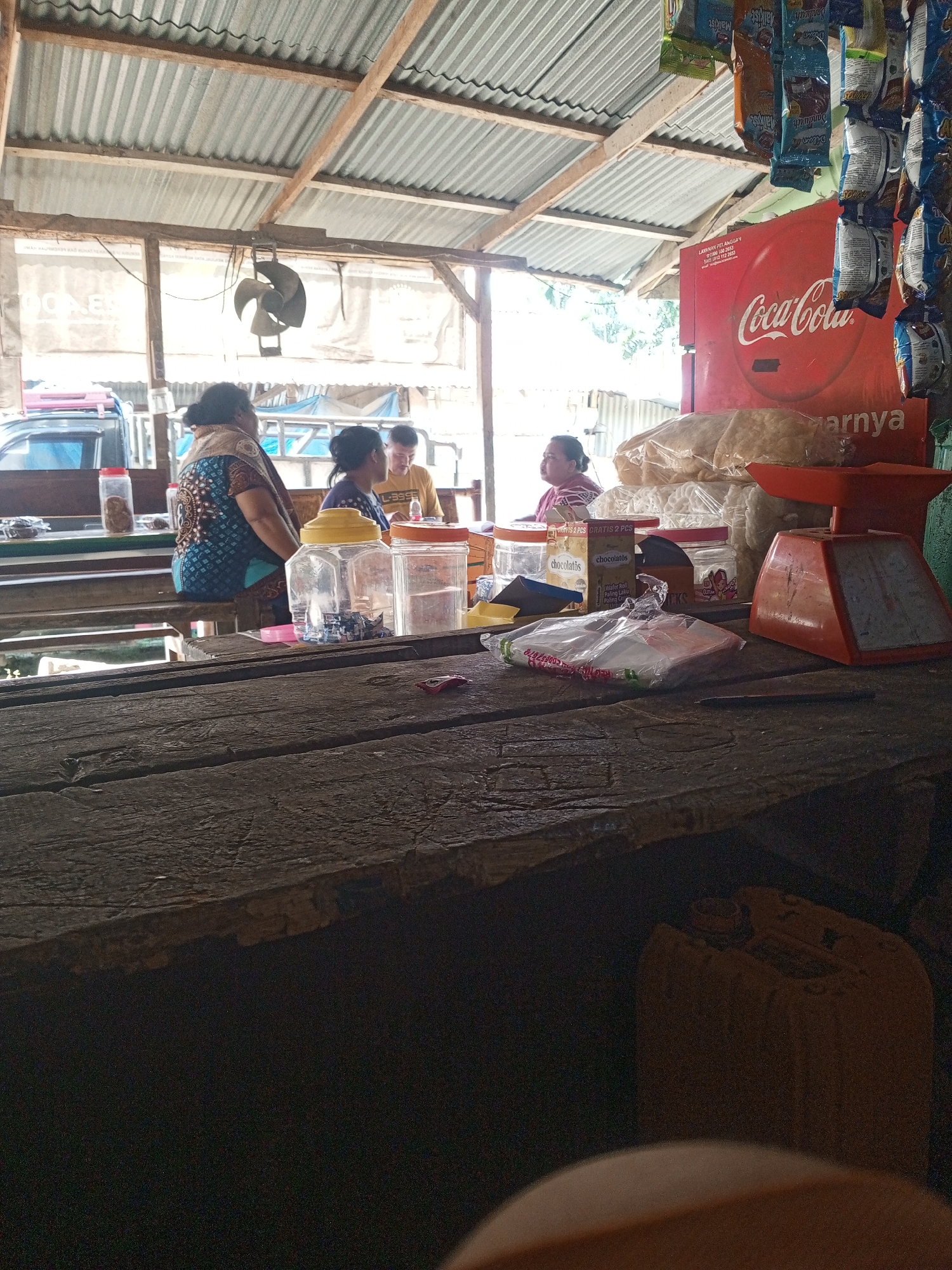 The image depicts an outdoor market stall in Tanah Gara Hulu, Indonesia. A corrugated metal roof supported by wooden beams covers the stall. In the background, a woman wearing a patterned blue and brown top sits on a stool, facing away from the camera. To her right, another person in a yellow shirt is visible. Various items are displayed on the wooden counter, including plastic containers with food items, a red Coca-Cola refrigerator, and a stack of bagged snacks. An orange weighing scale is placed on the counter. The foreground is dominated by a rough-hewn wooden table. The overall impression is of a simple, functional market environment.