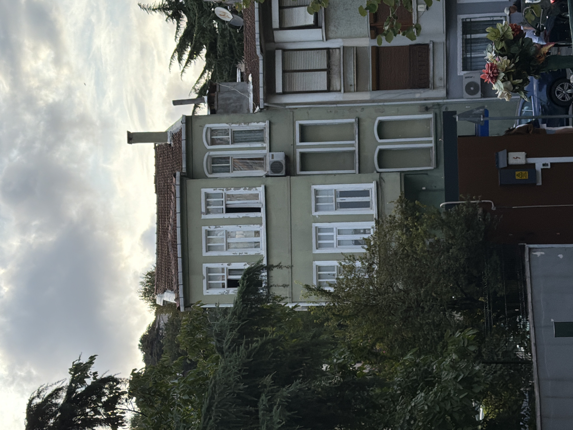 The image captures a close-up view of a narrow, multi-story building with a light green facade, located on a city street in Üsküdar, Turkey. The building features several white-framed windows, some of which appear to be open, revealing drawn curtains inside. A portion of a tiled roof is visible at the top, along with a chimney. Adjacent to this building, another structure with a lighter gray facade and brown shutters is visible. The foreground is occupied by lush green trees and foliage, obscuring the lower parts of the buildings. Further down the street, a dark blue car is parked, and two individuals are partially visible, one standing near the car and another walking away. A bouquet of artificial flowers in a dark green container is placed in the lower right corner of the frame. Electrical boxes and a warning sign are mounted on a brown structure in the lower left. The sky above is overcast with soft, diffused light, suggesting it might be late afternoon or early evening.