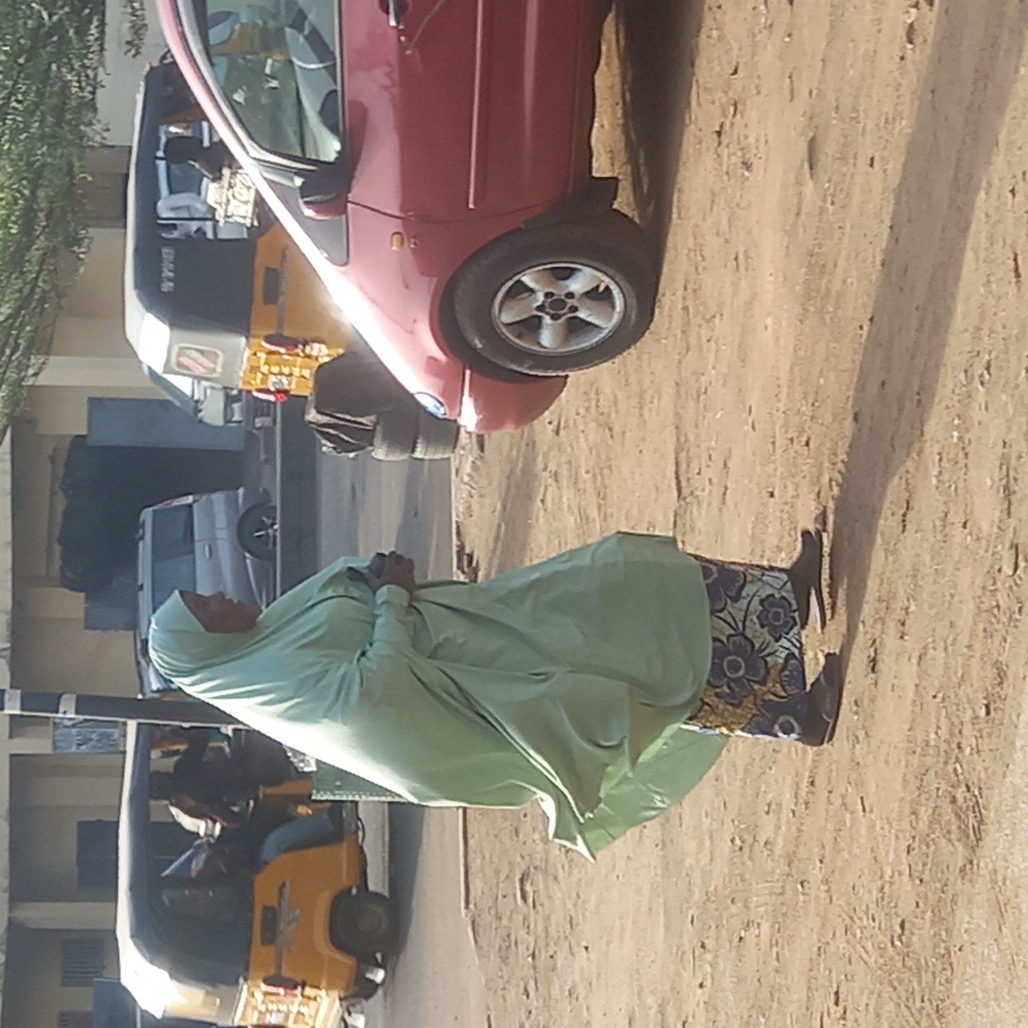 The image, taken in Dadi Riba, Nigeria, captures a sunlit street scene. In the foreground, a person stands on the right side of an unpaved, sandy road. They are dressed in a flowing, light green outer garment that covers most of their body, including a headscarf (hijab or niqab). The lower part of their attire reveals a patterned skirt or dress in dark blue with light-colored floral motifs. Their hands are clasped together at the front. Long shadows stretch across the sandy ground, suggesting either early morning or late afternoon sunlight.
Behind the person, to their right, a red car, possibly a hatchback, is parked or stopped. It has silver multi-spoke wheel rims. Further down the street, on the left side, two yellow and black auto-rickshaws (often called Keke Napep in Nigeria) are visible, one more prominently in the midground and another further back. A white sedan is also parked or moving in the distant background on the left, near some light-colored buildings and green foliage. The scene appears to depict everyday life on a dusty road in a residential or commercial area. No legible text is visible in the image.