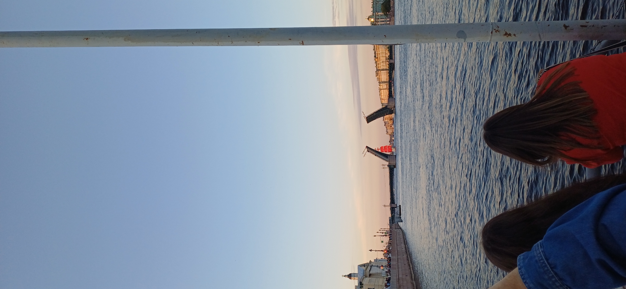 The image captures a scene on a body of water, likely a river or canal, in Saint Petersburg, Russia, during what appears to be either dawn or dusk due to the soft, warm light. In the foreground, two individuals are seen from behind, their backs to the camera, looking out towards the water. One person wears a blue denim jacket, and the other a bright orange top.
Dominating the middle ground is a bascule bridge, opened in the center, allowing passage for a vessel. The bridge is flanked by buildings lining the waterfront on both sides of the water. A prominent, bright red schooner with sails furled is positioned beneath the opened bridge, its vibrant color a striking contrast against the muted tones of the cityscape and the water. The sky is a clear gradient of pale blue and soft pinkish-orange, suggesting twilight. On the left bank, there are several people walking along the embankment, with an ornate building featuring a dome visible. On the right bank, grand classical buildings with multiple windows and architectural details are visible. A tall, weathered white pole stands in the right foreground, partially obscuring the view. The water is a deep blue with visible ripples, reflecting the light from the sky.