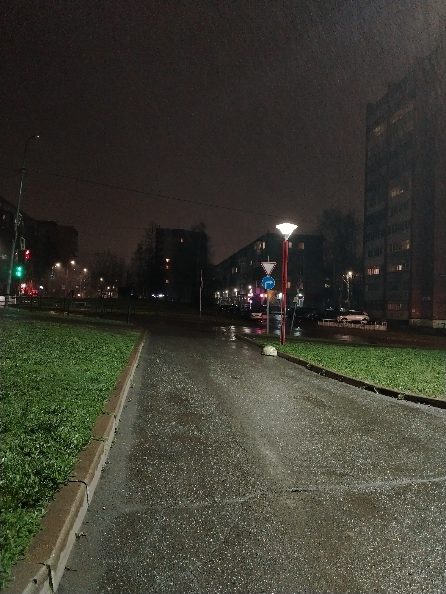 The image captures a street scene in Izhevsk, Russia, at night, likely during or after rainfall, as the pavement and surfaces appear wet. The foreground is dominated by an asphalt path, and a strip of green grass borders the path with a concrete curb. Further down the street, a lamp post with a round light sits next to road signs. Parked cars line the side of the road, and tall residential buildings are visible in the background. The sky is dark, and the overall lighting is dim. A traffic light shows green on the left side of the frame.